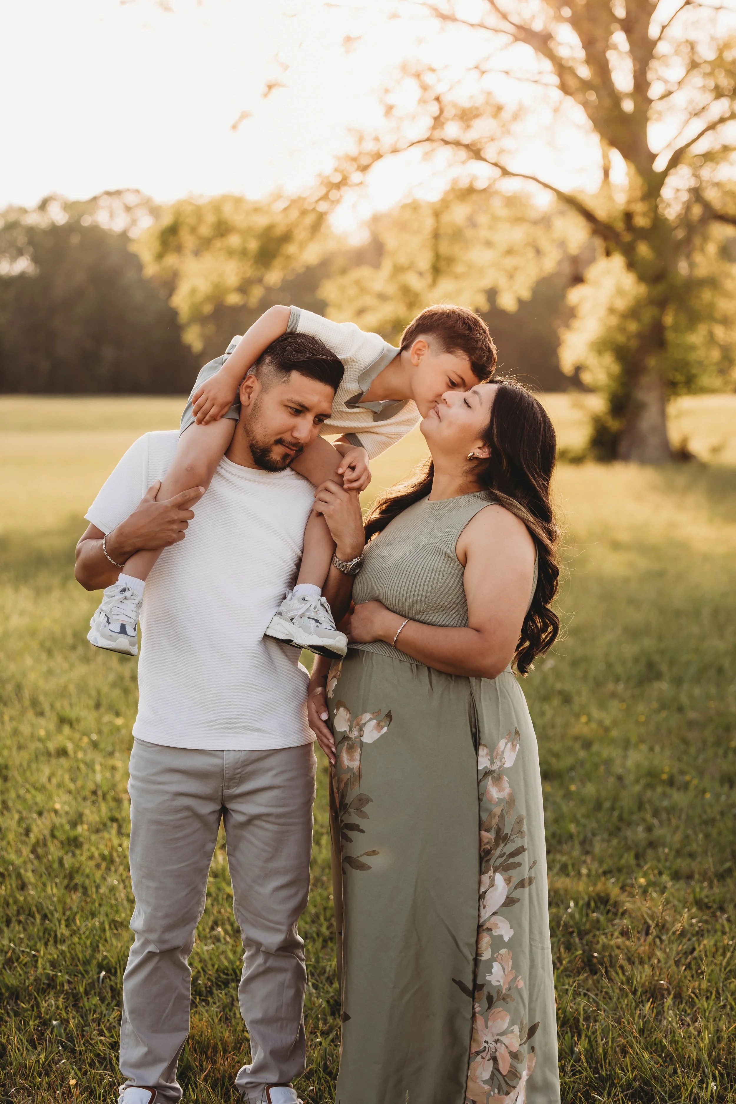 A family of three outdoors during sunset, with the father carrying the son on his shoulders and the mother standing close, all sharing a tender moment in a park surrounded by green grass and trees.