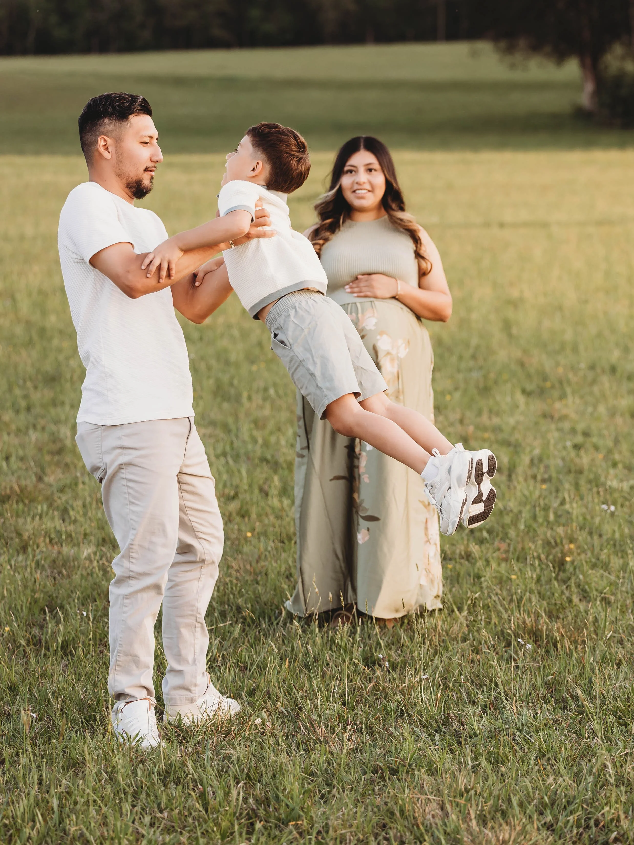 A man and woman standing in a grassy field with a young boy. The man is holding the boy, who is wearing a white shirt and beige shorts, while the woman stands nearby with one hand on her stomach. The background features a large, open grassy area and 