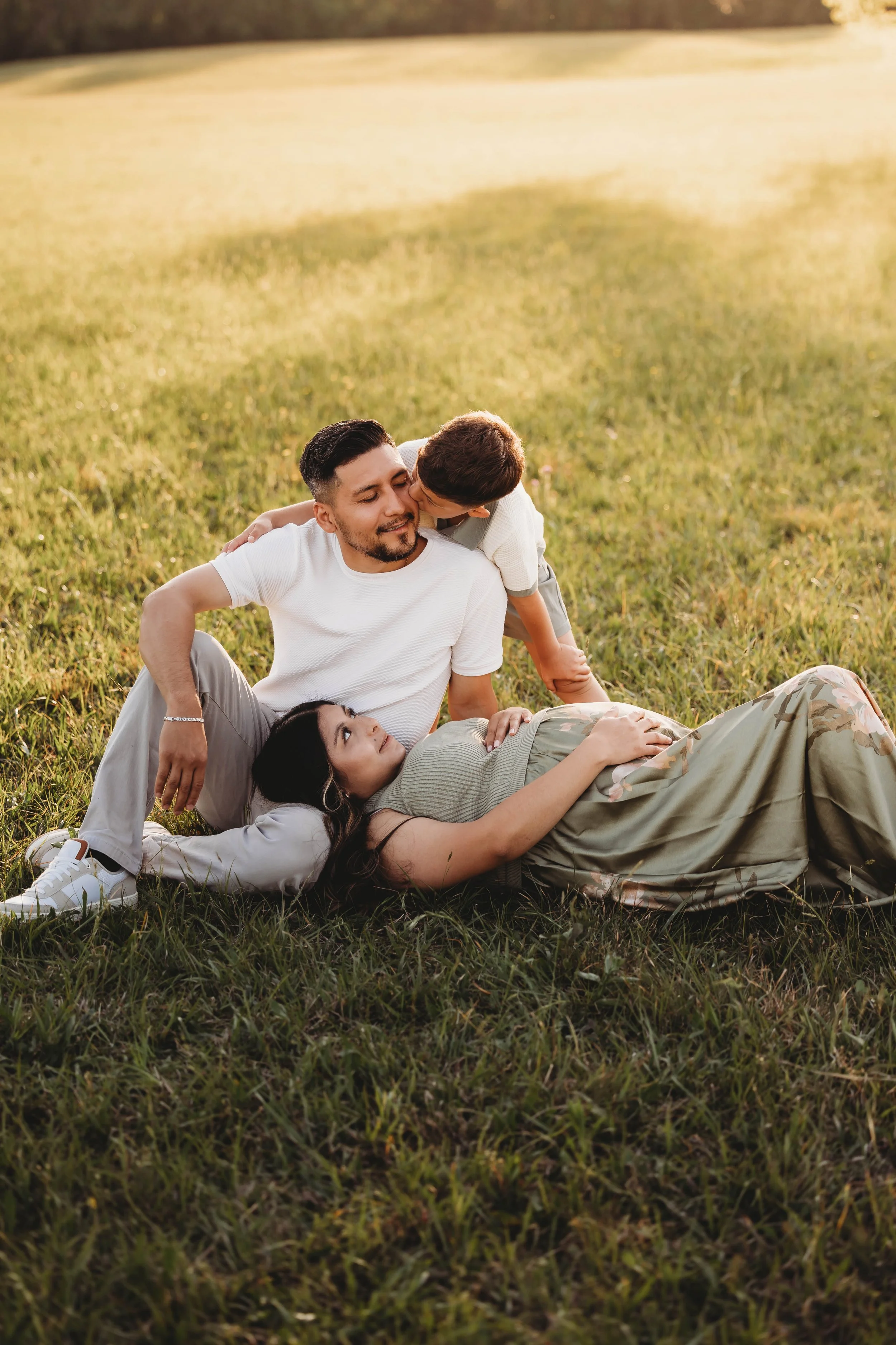 A family spending time together outdoors on a grassy field during sunset, including a man, woman, and a young boy, with the boy giving the man a kiss on the cheek.