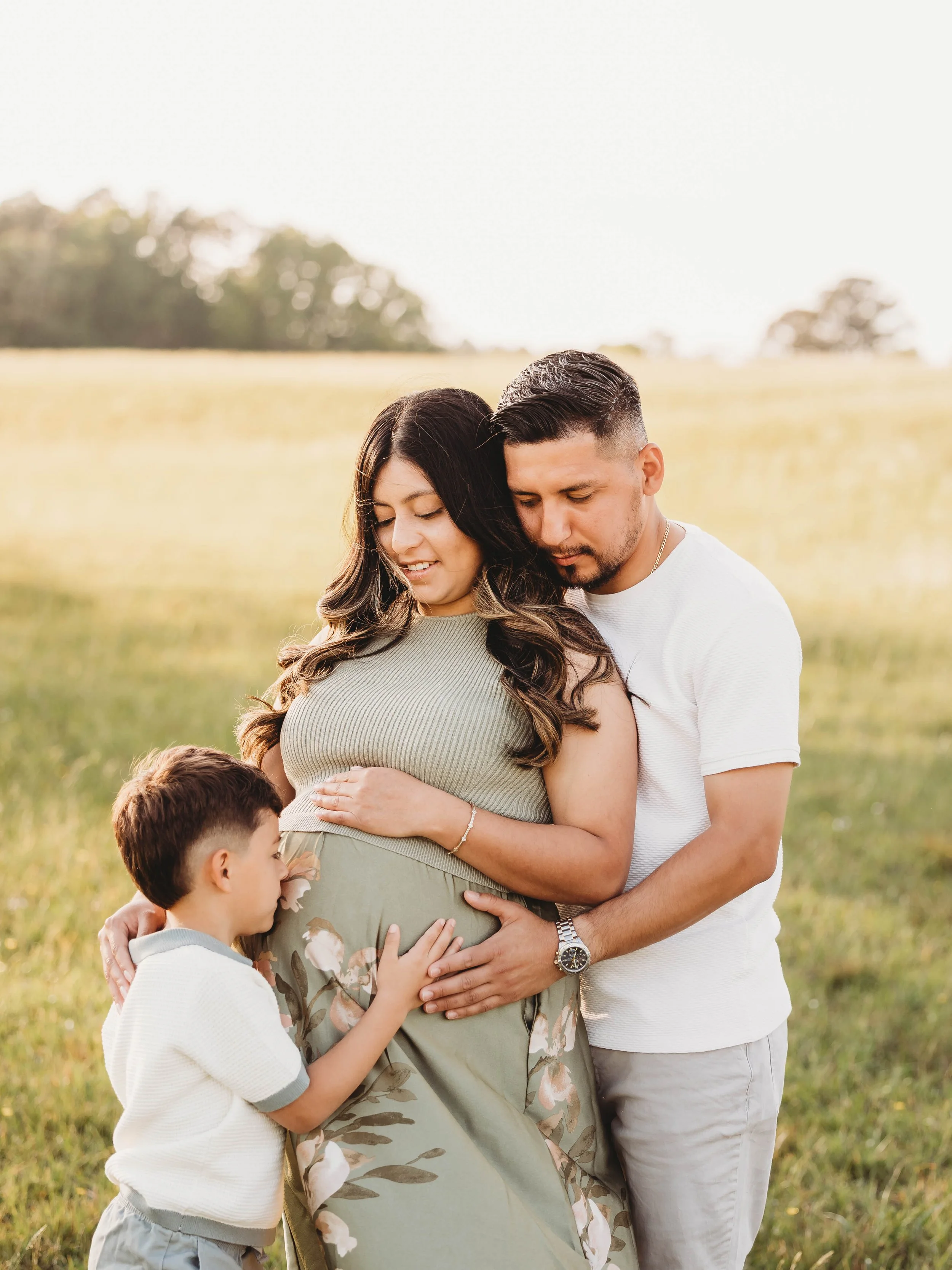 A pregnant woman in a floral dress is embraced by a man, with two children touching her belly, in a grassy field during daylight.