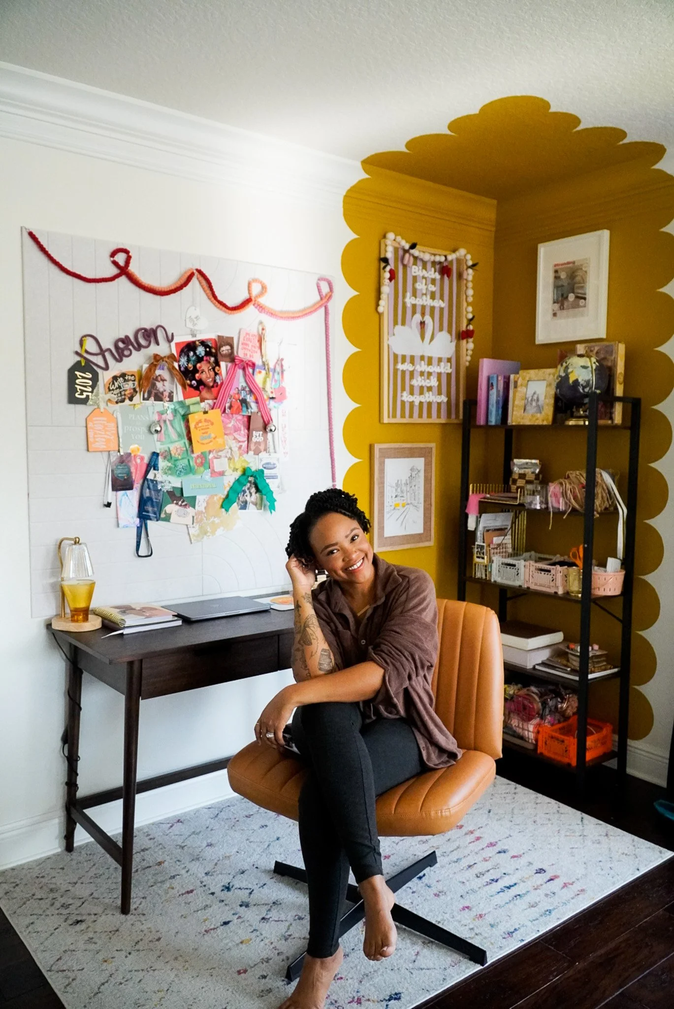 A woman with tattoos sitting on a tan office chair in front of a desk, smiling at the camera. The room has a white wall with a board decorated with photos and notes, a mustard-colored wall with scalloped edges, a black shelving unit with books and decor, and a framed picture on the wall.
