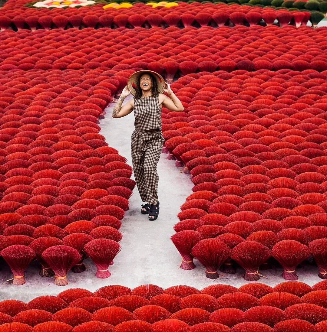 A woman smiling and laughing while walking through a path in a field of red dried flowers, wearing a checked sleeveless top, checked pants, sandals, and a straw hat.