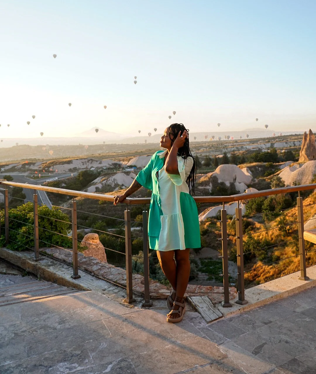 A woman standing on a balcony overlooking a landscape with rock formations and hot air balloons in the sky at sunrise or sunset.