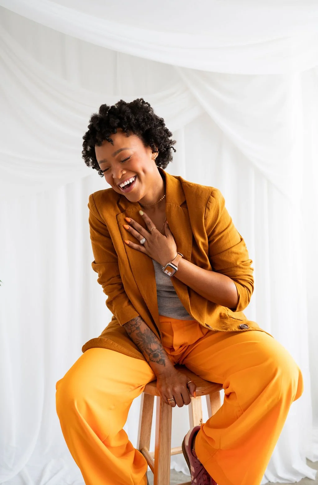 A woman  sitting on a wooden stool, laughing with her eyes closed and hand on her chest. She is wearing a mustard-colored blazer, matching pants, and a gray top, with a smartwatch, rings, and a tattoo on her arm. The background has white draped fabric.