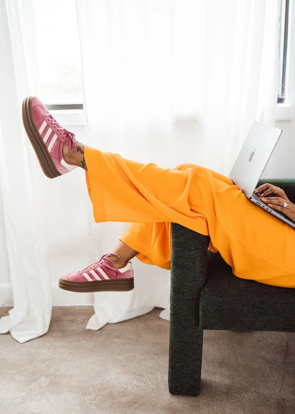 Person relaxing on a dark gray couch with legs up, wearing bright orange pants and pink Adidas sneakers, using a silver laptop in a well-lit room with white curtains and a window behind.