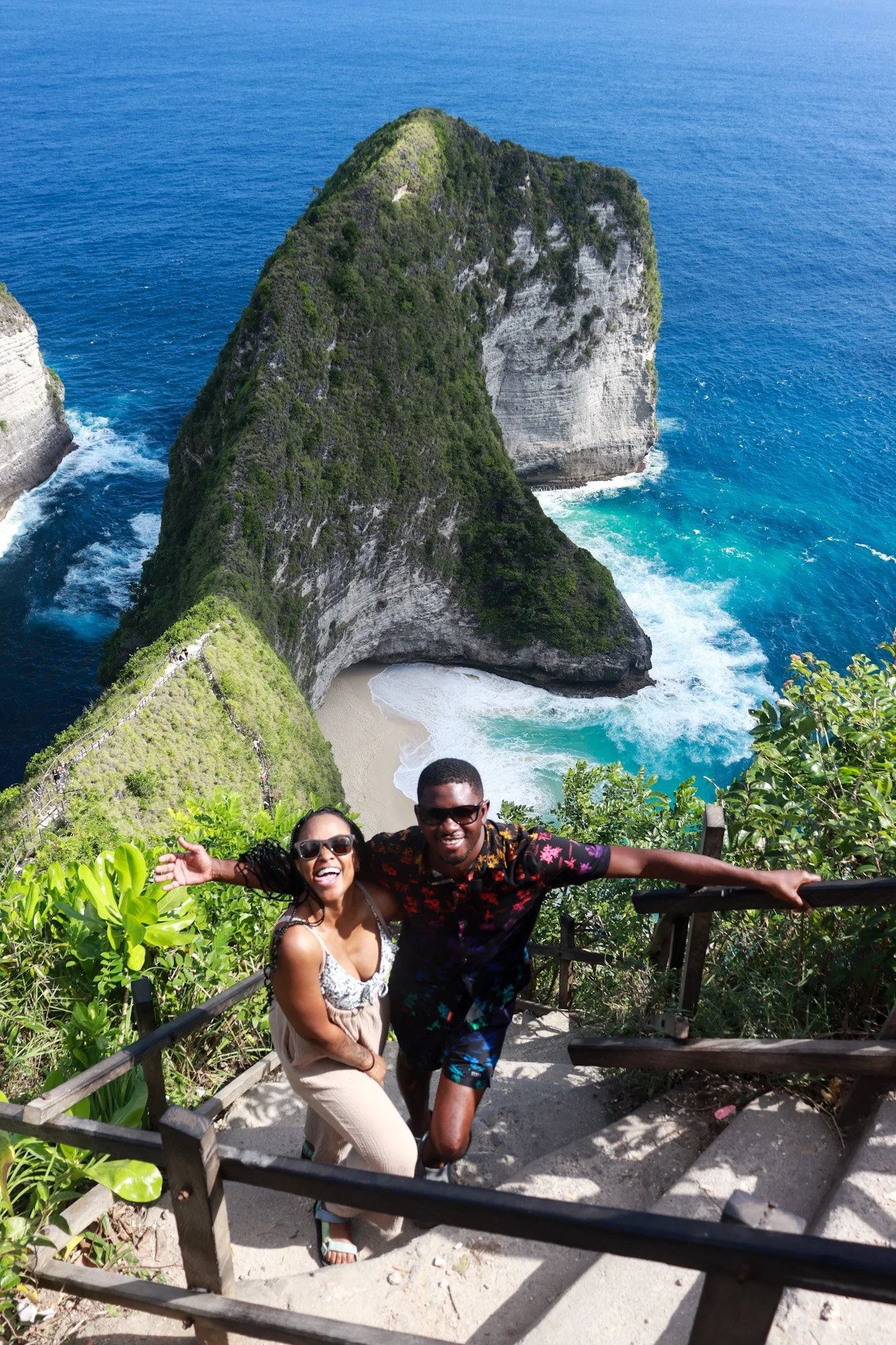 Two people smiling and posing on a staircase with lush green foliage, overlooking a scenic view of a beach with a large rock formation in the ocean and a cliff in the background.