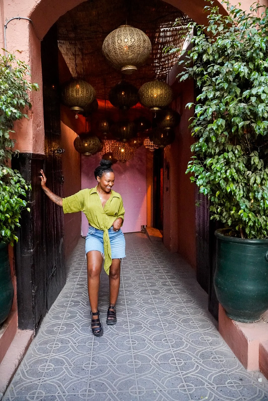 A woman in a green shirt and denim shorts standing in a decorated archway, surrounded by hanging pendant lights and potted plants.