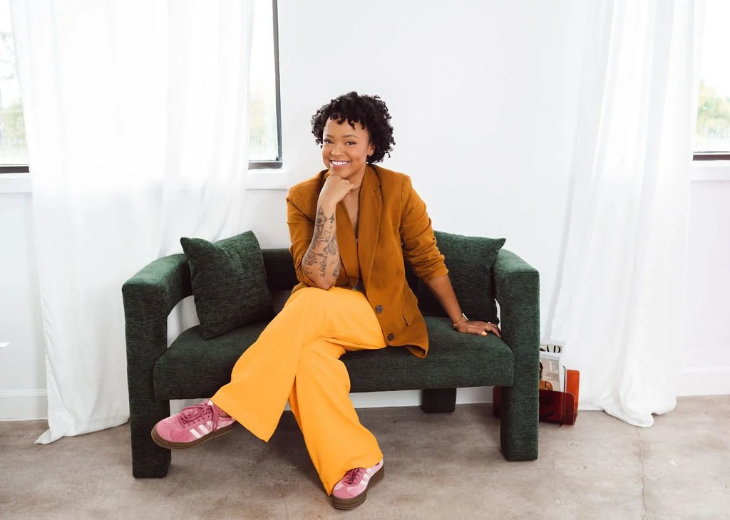A woman with curly hair and tattoos smiling while sitting on a dark green couch in a bright room with white curtains and windows.