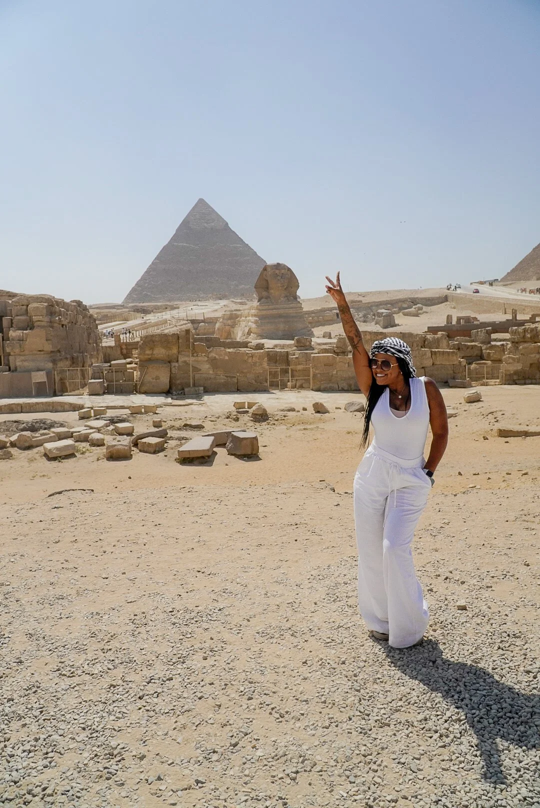 A woman dressed in white with sunglasses and a headscarf standing in front of the Great Pyramid and the Sphinx in Egypt, making a peace sign with her hand.