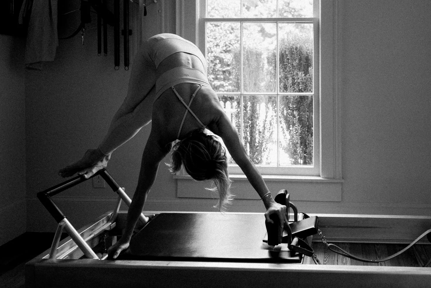A woman in workout attire performing a yoga or stretching pose on a reformer machine in a room with a large window, sunlight shining through the window, and various exercise tools hanging on the wall.