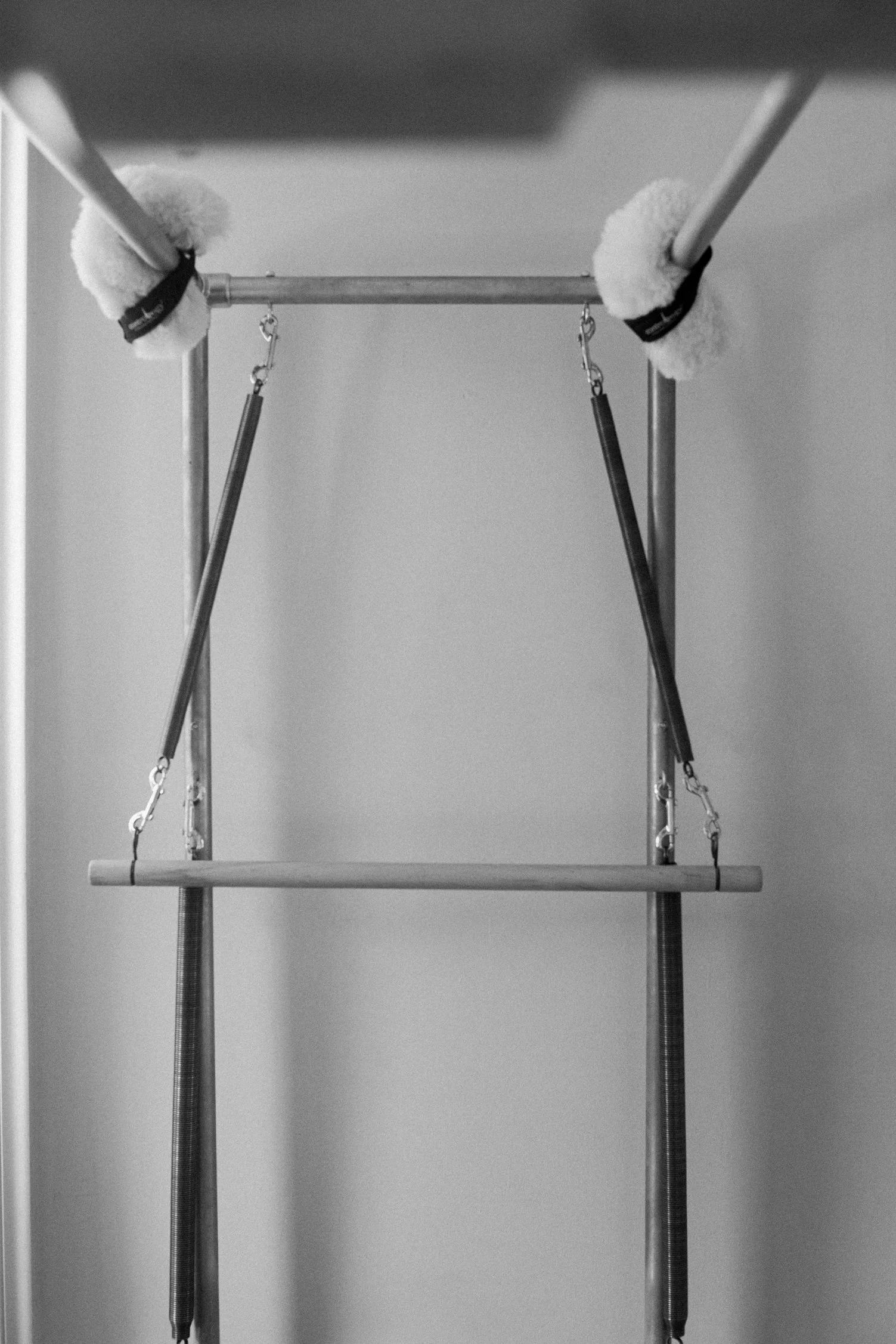 A black and white photo of a baby jumper with a wooden seat, secured to an overhead bar with elastic straps and plush cushions at the top.