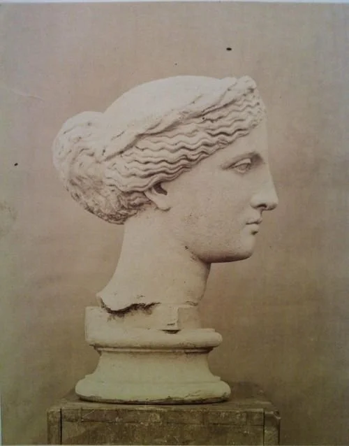 A bust of a woman from ancient Greece or Rome, made of stone, showcasing detailed wavy hair, a serene expression, and classical features.