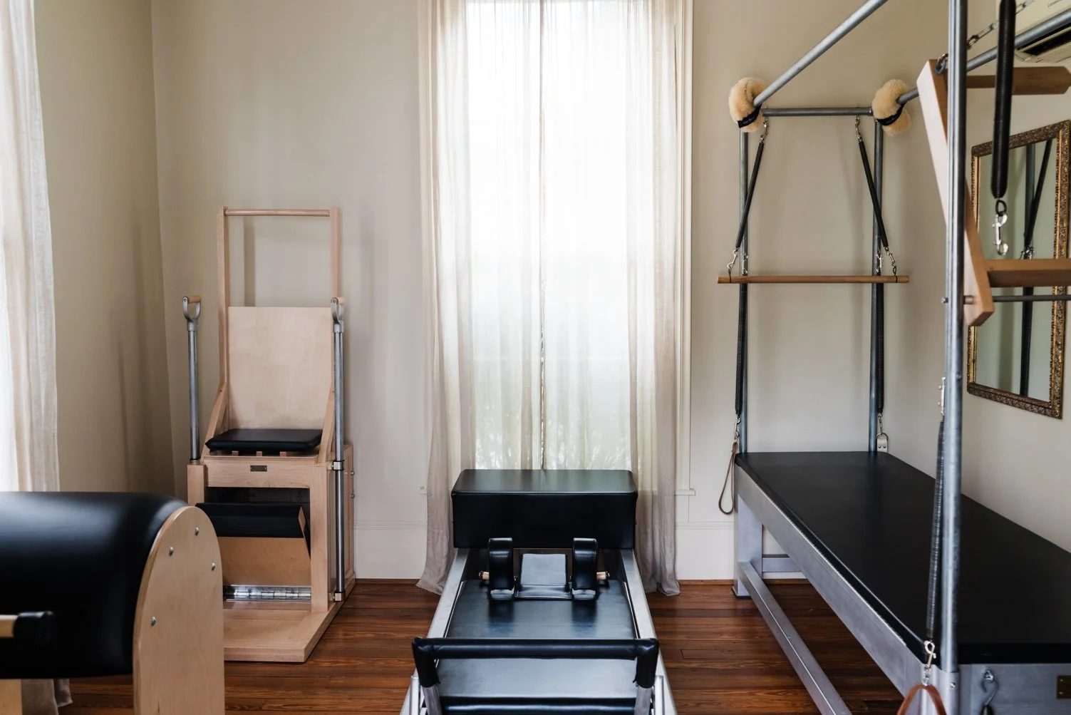 Room with Pilates equipment, including a Pilates chair, a ladder barrel, and a Cadillac, with a window and mirror on the wall.