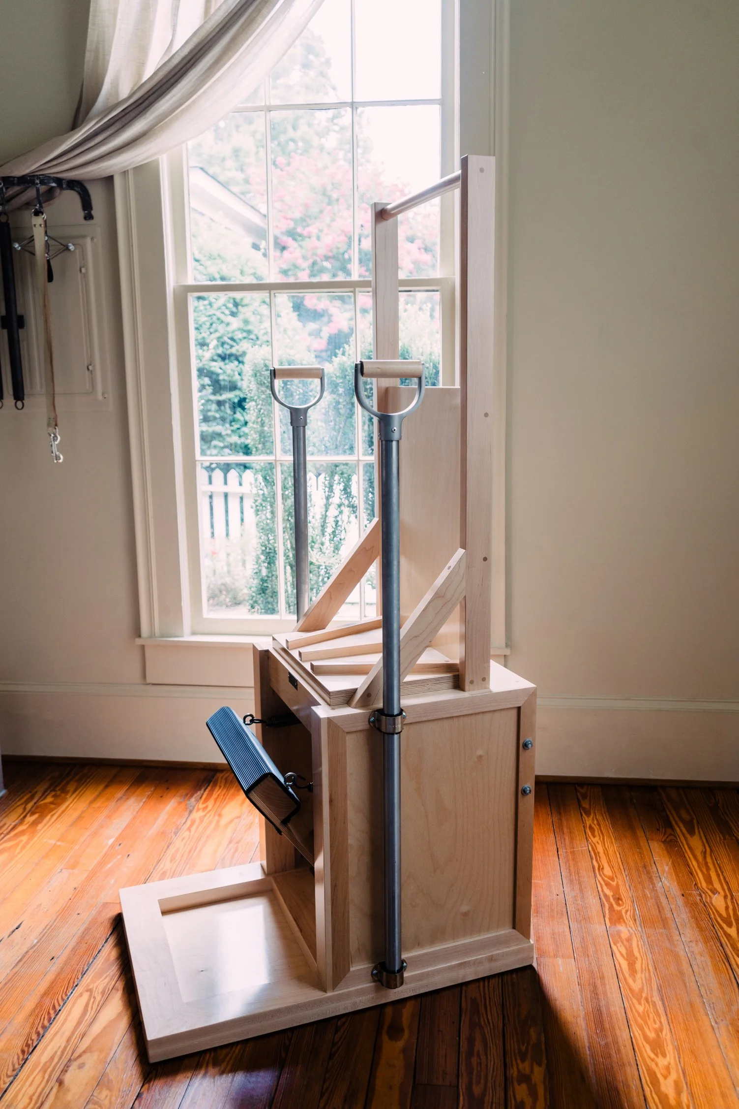 Unfinished wooden climbing structure with metal handrails by a window with white curtains, in a room with wooden flooring.