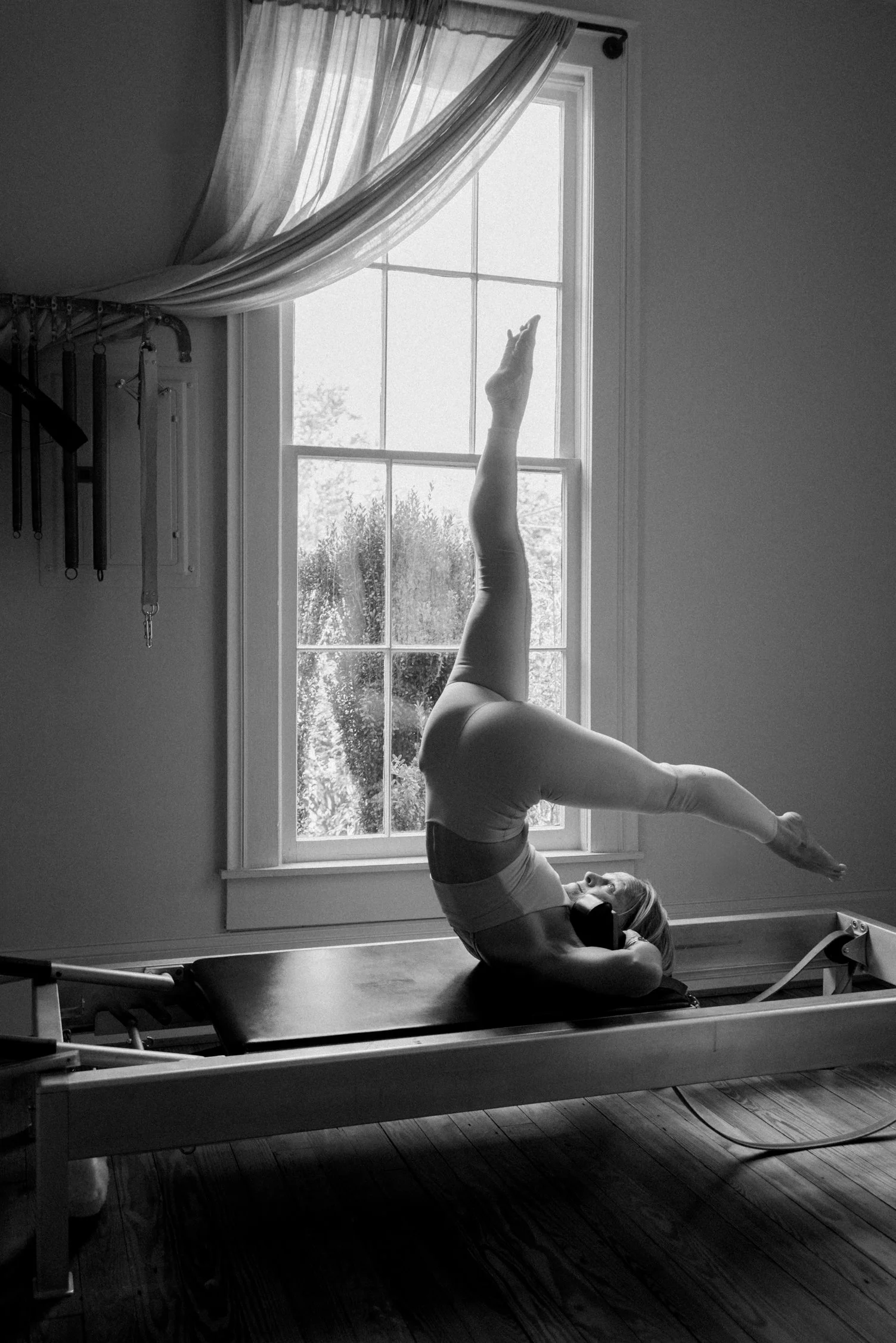 A woman practicing aerial yoga on a reformer machine in a room with a large window and curtains, black and white photo.