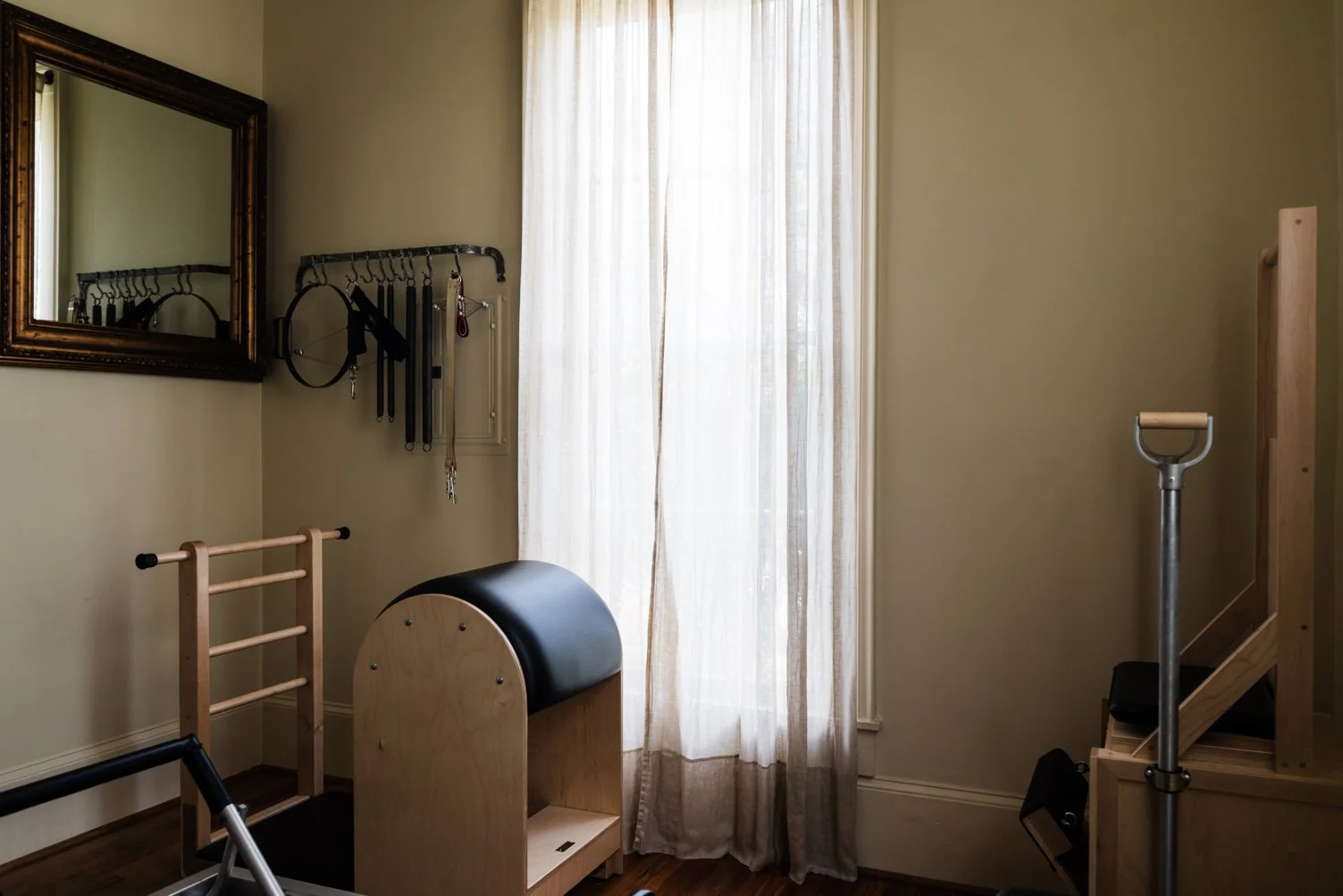 A room with fitness equipment including a wooden Pilates barrel, a wooden dashboard, a balance beam, and a small step on a staircase. There's a wall-mounted rack holding resistance bands and other gym accessories, a mirror, and a window with sheer curtains.