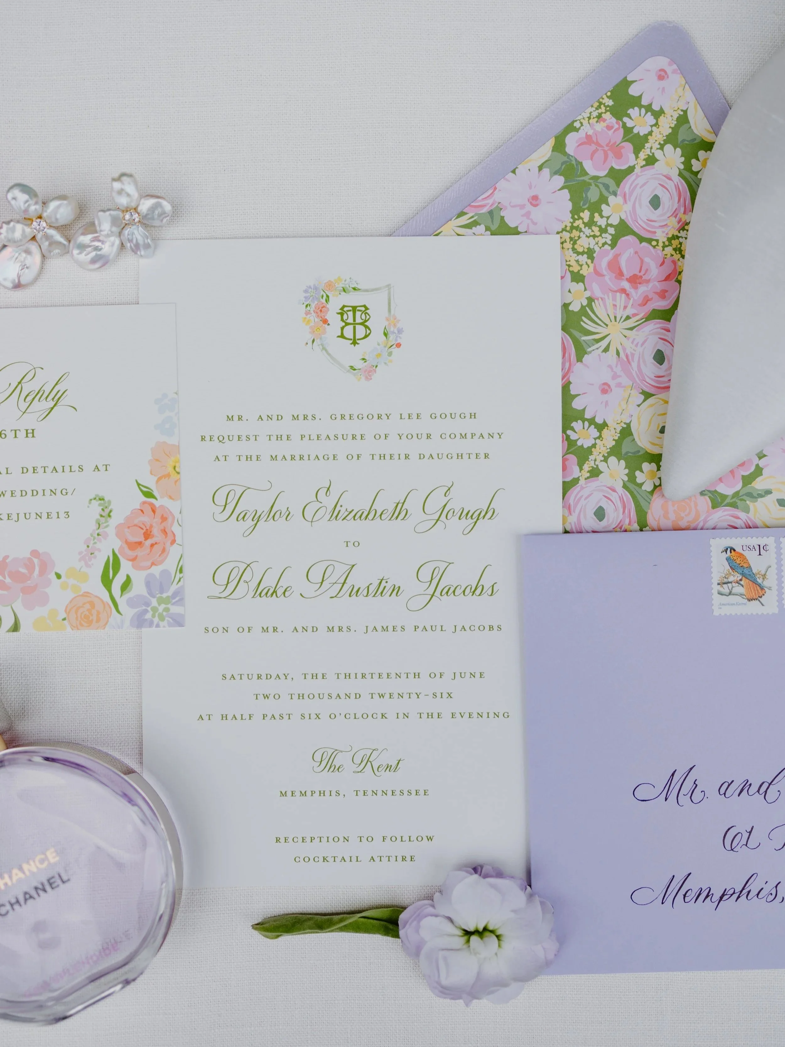 A wedding invitation set with floral design and monogram, surrounded by decorative flowers, an envelope with a stamp, and a small flower, on a white background.