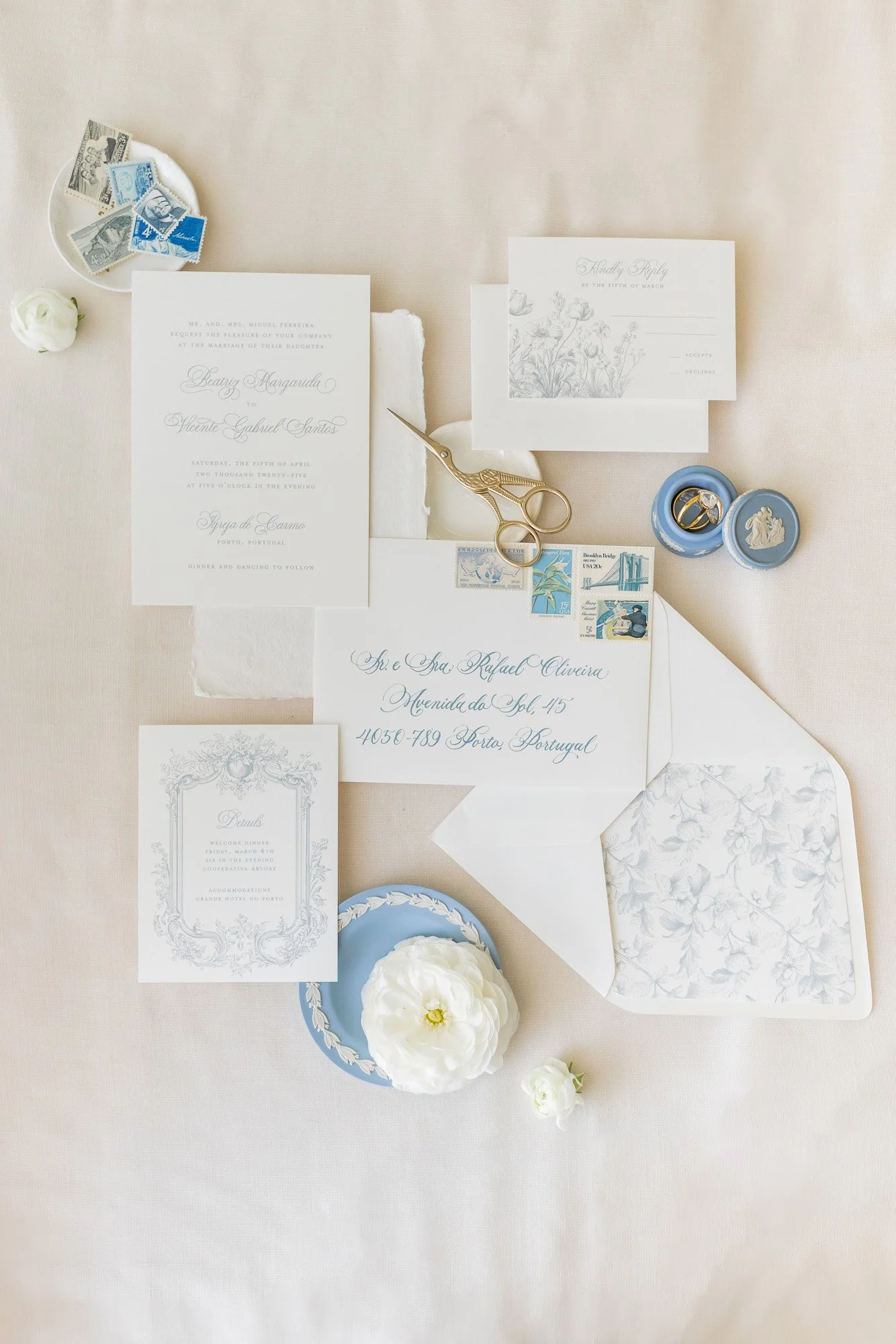 A flat lay photograph of wedding stationery on a beige tablecloth, including an invitation, RSVP card, envelope with stamps, a pair of gold scissors, a white flower, two small blue containers with rings, and a small dish with vintage postage stamps.
