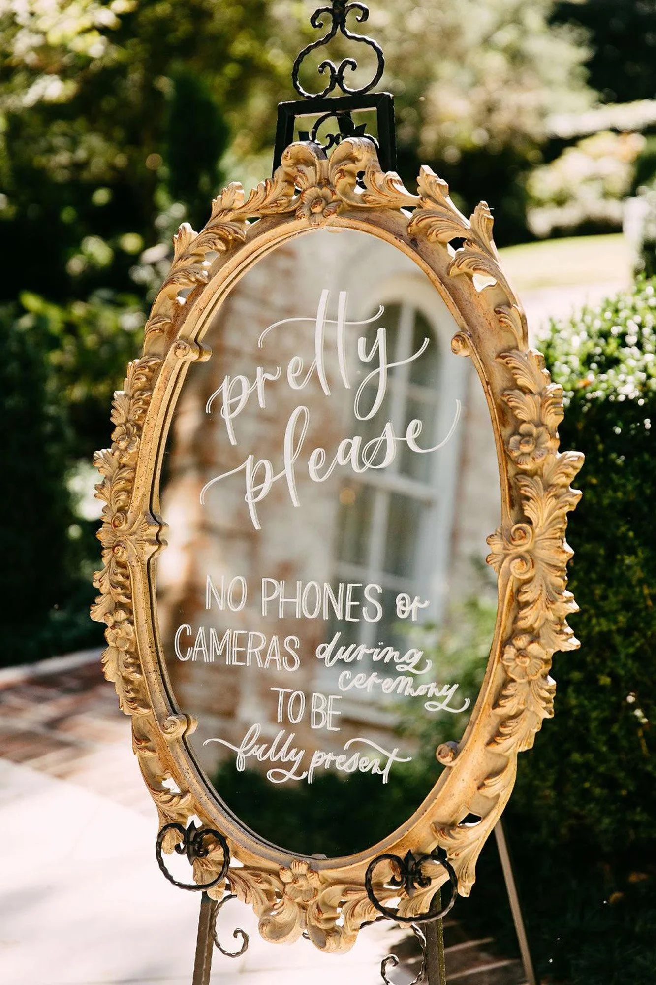 A decorative gold-framed mirror with a sign written on it in white cursive and block text that reads, 'pretty please NO PHONES or CAMERAS during ceremony TO BE fully present,' outside in a garden area with green bushes and a brick-building reflection