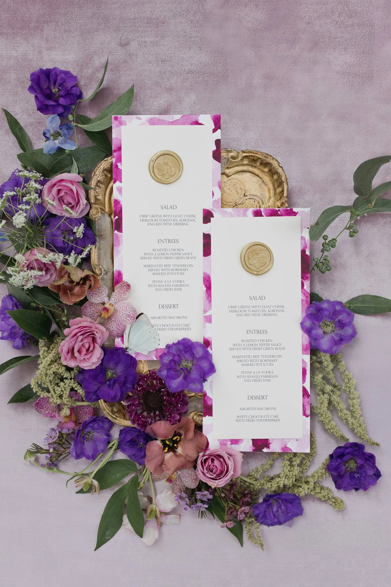 Elegant wedding menu cards surrounded by purple, pink, and white flowers with greenery, laid on a pink fabric background.
