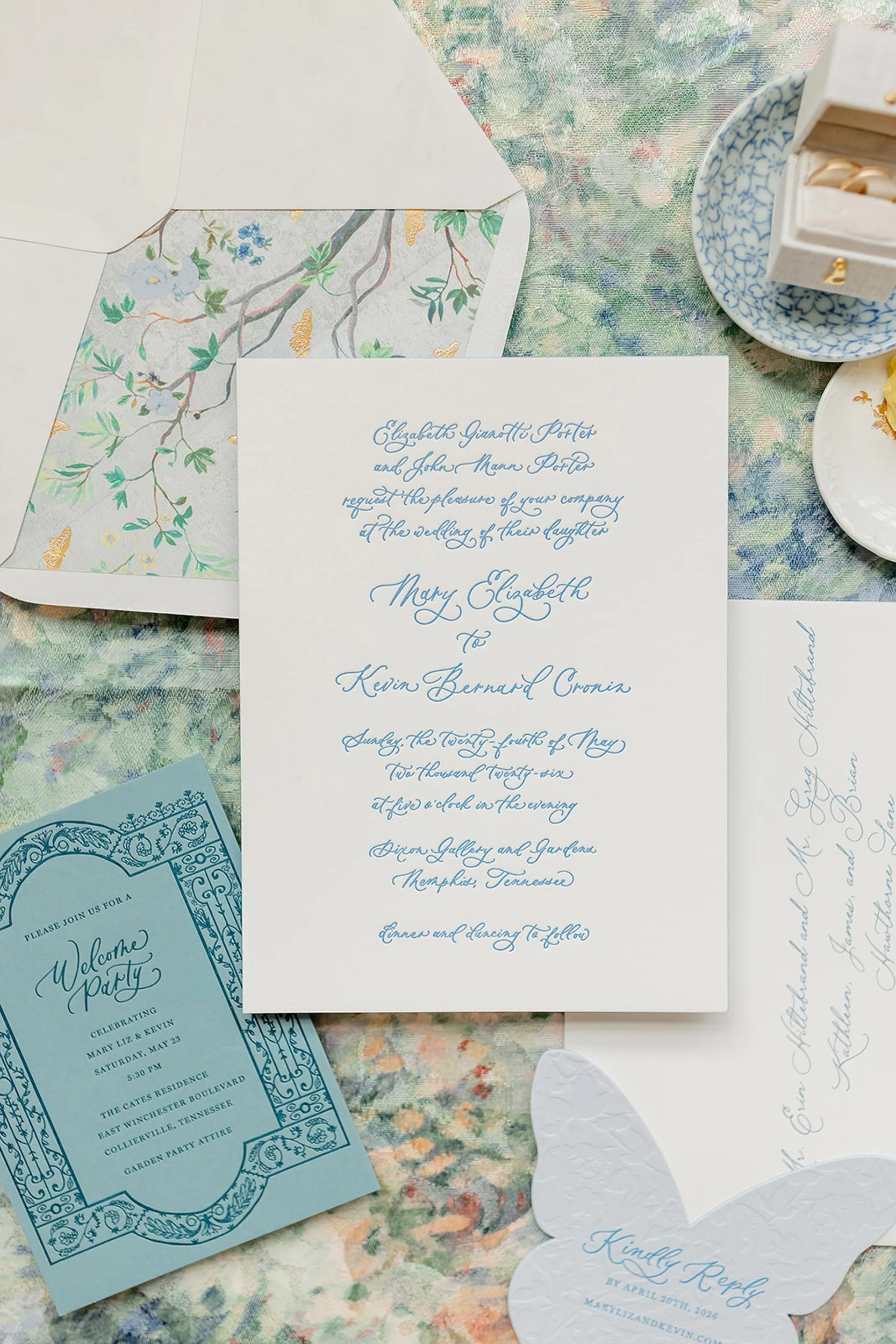 A collection of wedding invitations and event stationery on a colorful textured background, including a white invite with blue cursive writing, a blue event card, a blue and white patterned decorative dish, and a white heart-shaped paper with blue cursive text. Some of the stationary features floral and decorative designs.