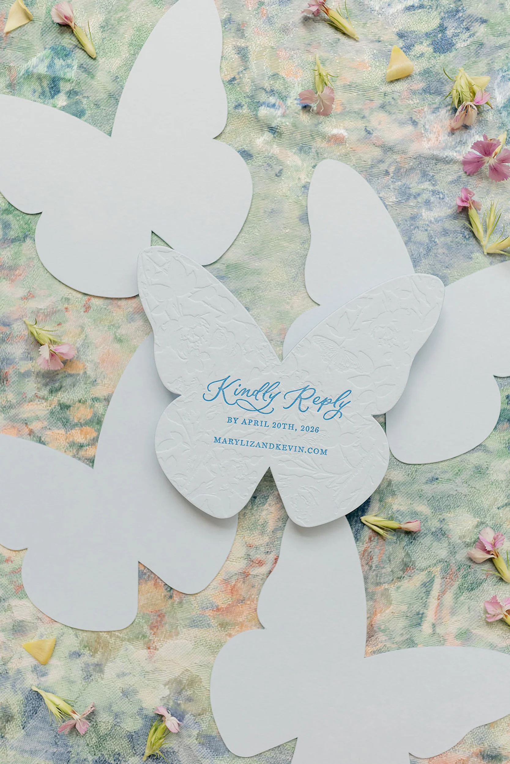 White butterfly-shaped paper cutouts with embossed patterns on a pastel-colored fabric background, scattered with small pink and yellow flower petals. One cutout has blue text reading, 'Kindly Reply by April 20th, 2026 Marylandkevin.com'.