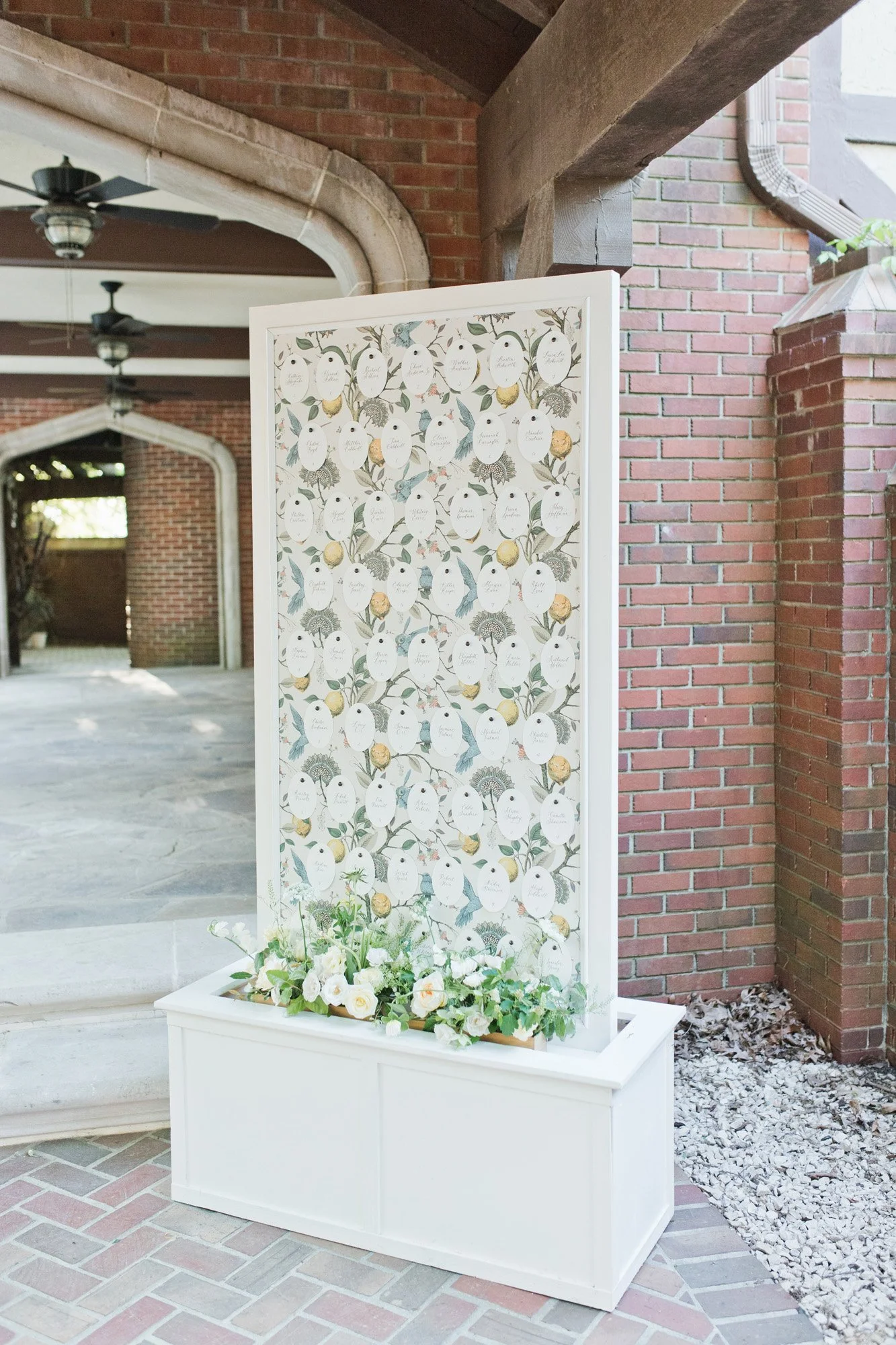 Decorative wedding or event seating chart with flower and bird designs in a white frame and base, featuring white oval tags with handwritten names hanging on a floral and bird pattern background.
