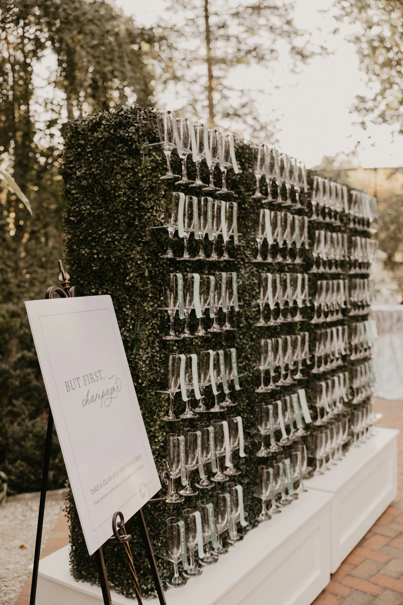 A wall display of champagne glasses hanging upside down with a sign that reads 'But First... Champagne' at an outdoor event.