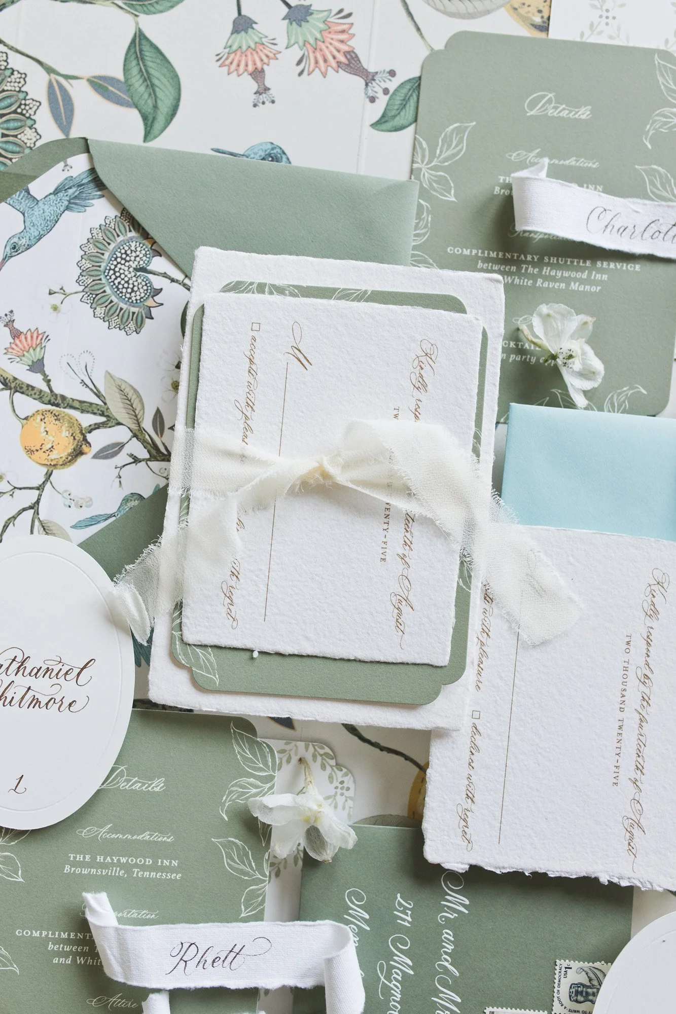 A collection of wedding stationery including invitations, RSVP cards, and envelopes in pastel green, blue, and white, adorned with floral and botanical designs, with some pieces tied with white ribbon and decorated with small white flowers.