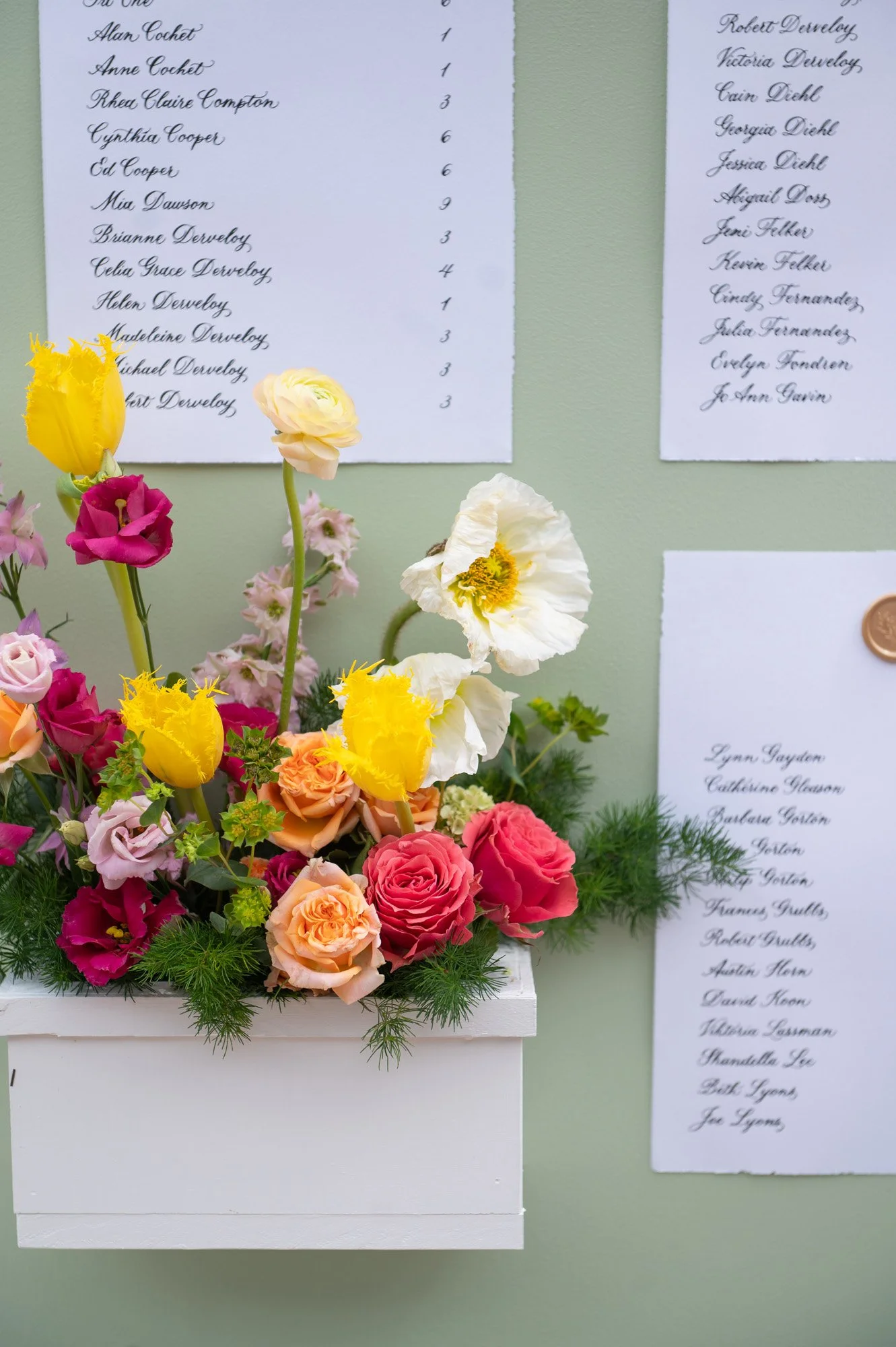 A white flower arrangement in a white box with various colorful flowers including yellow tulips, pink roses, and white poppies, is placed against a light green wall with handwritten lists on white paper attached with a copper seal.