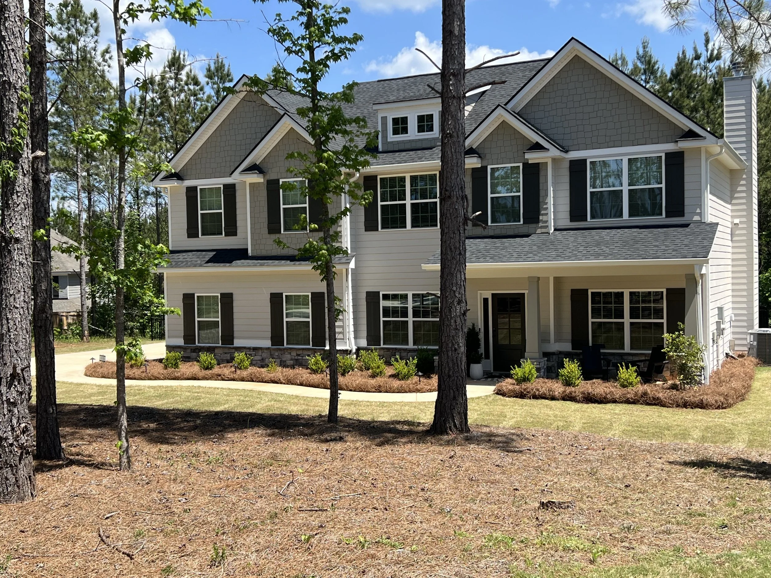 A white three-story house with black shutters and a gray roof partially obscured by trees, with a well-maintained lawn and landscaped garden in front and a bright blue sky with clouds overhead.