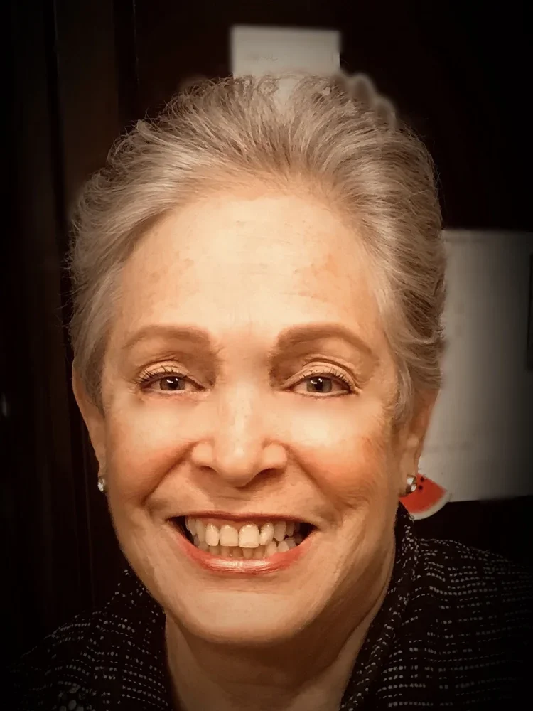 Close-up of a smiling Linda with short gray hair, wearing earrings and a dark, patterned top, indoors.