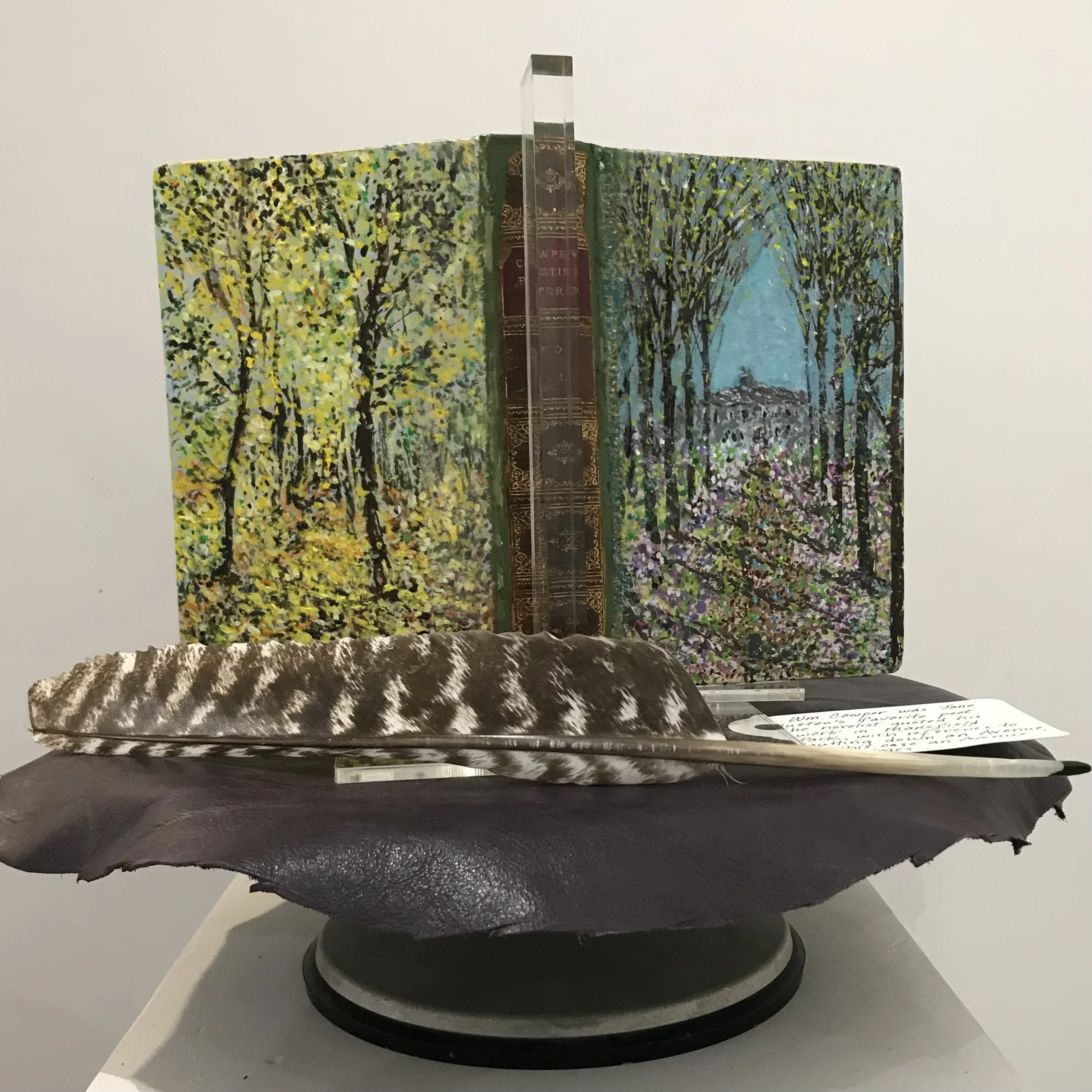 A painted book with a landscape scene of trees and a blue sky, displayed on a stand. A feather with a black and white pattern and a handwritten note are in front of the book.