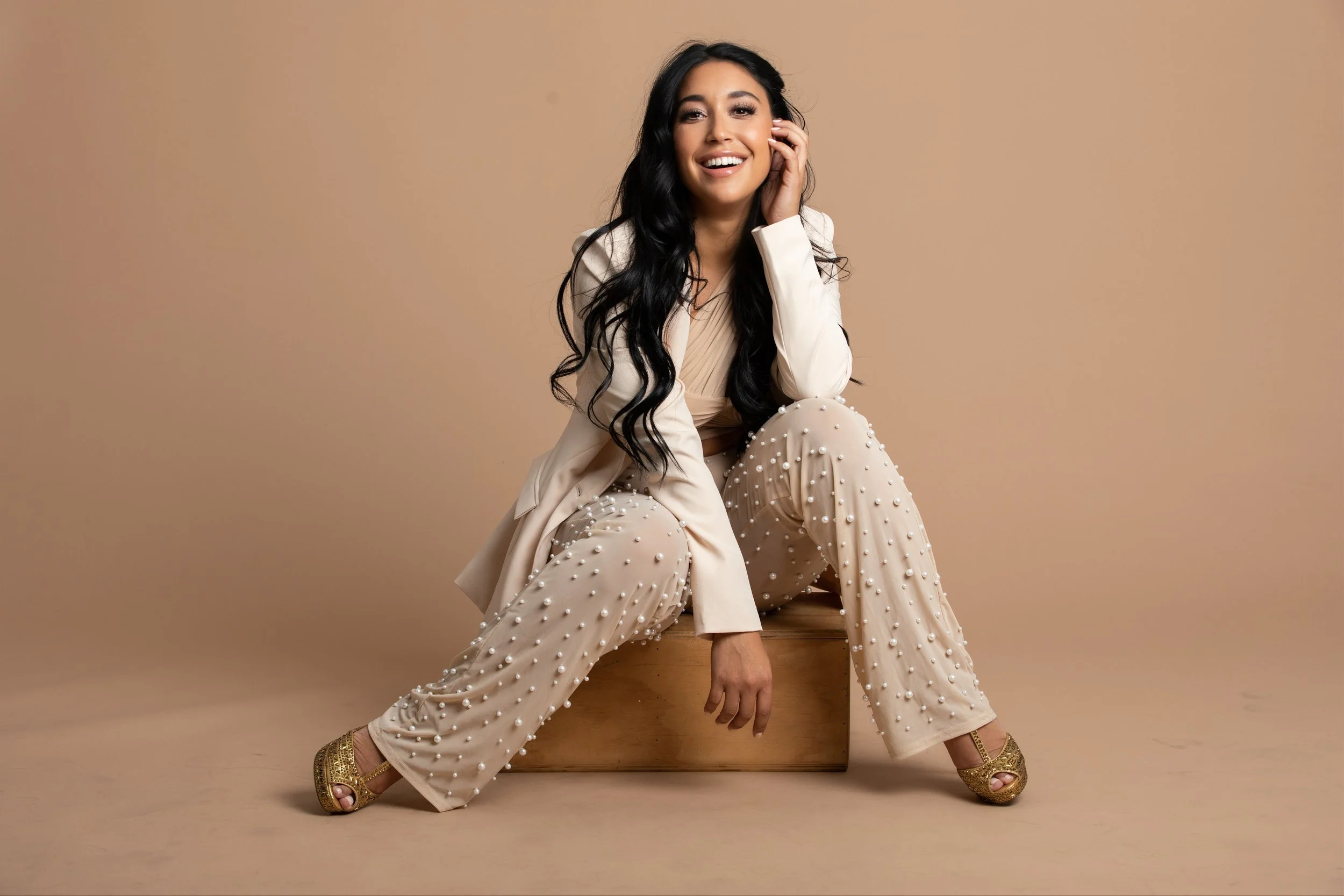 A woman with long black hair, wearing a beige suit and pearl-embellished pants, sitting on a wooden box with a beige backdrop, smiling and touching her face.