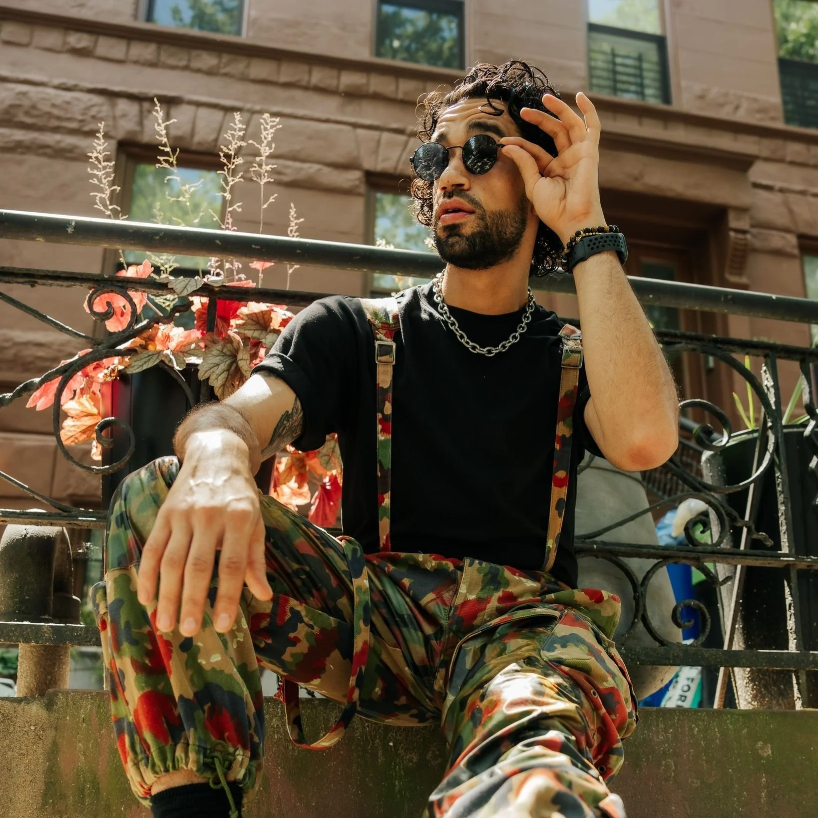 A man with curly hair, sunglasses, black t-shirt, camouflage cargo pants, and accessories sitting on outdoor steps, with a building and plants in the background.