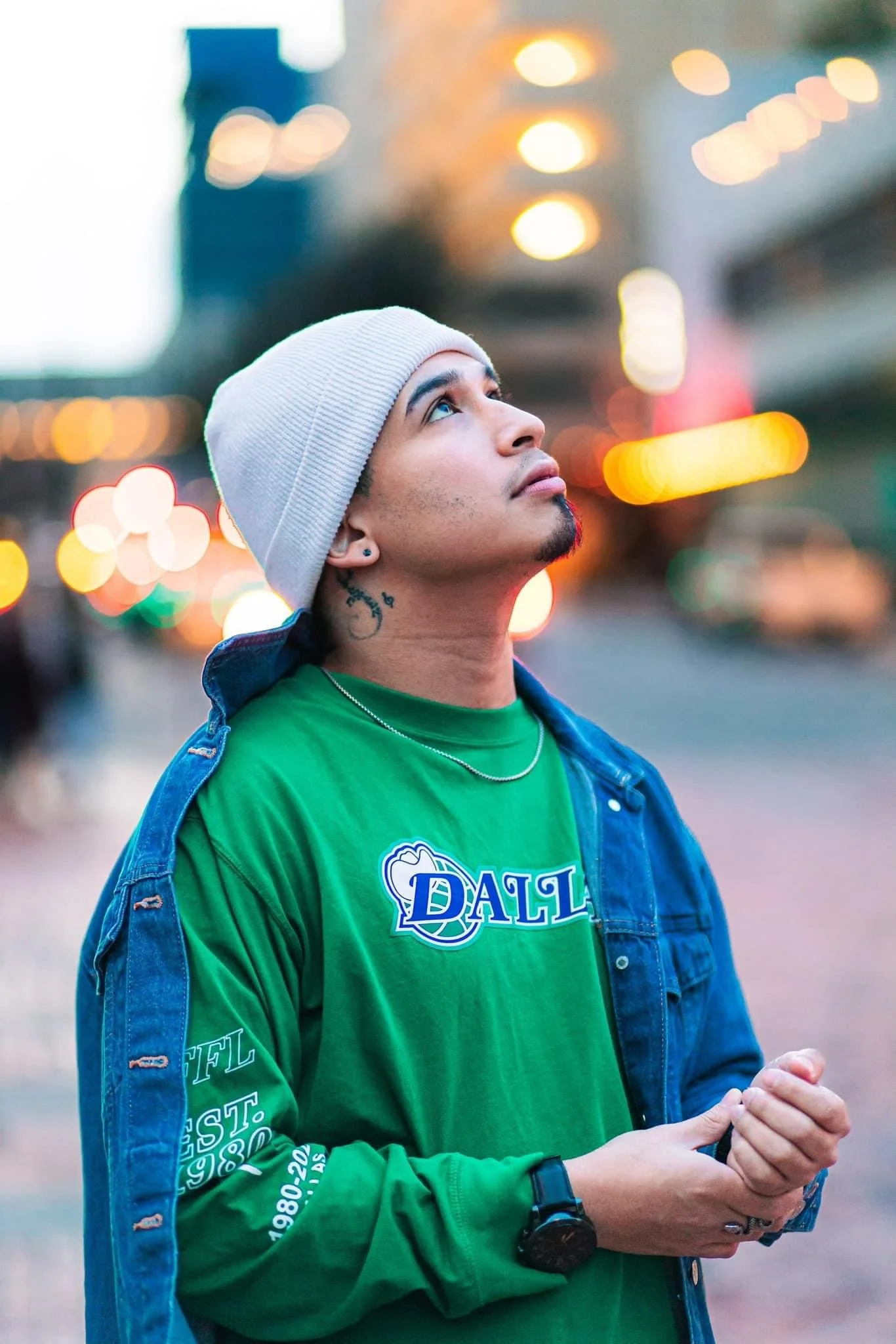 A young man with a white beanie hat, piercing blue eyes, and tattoos on his neck, standing outdoors in an urban setting during evening, looking up, wearing a green T-shirt with text and a denim jacket, with blurred city lights in the background.