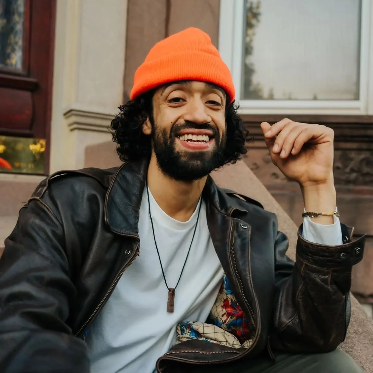 A smiling man with curly hair and a beard, wearing a bright orange beanie, black leather jacket, white t-shirt, and a necklace, sitting on steps outside a building with a window in the background.
