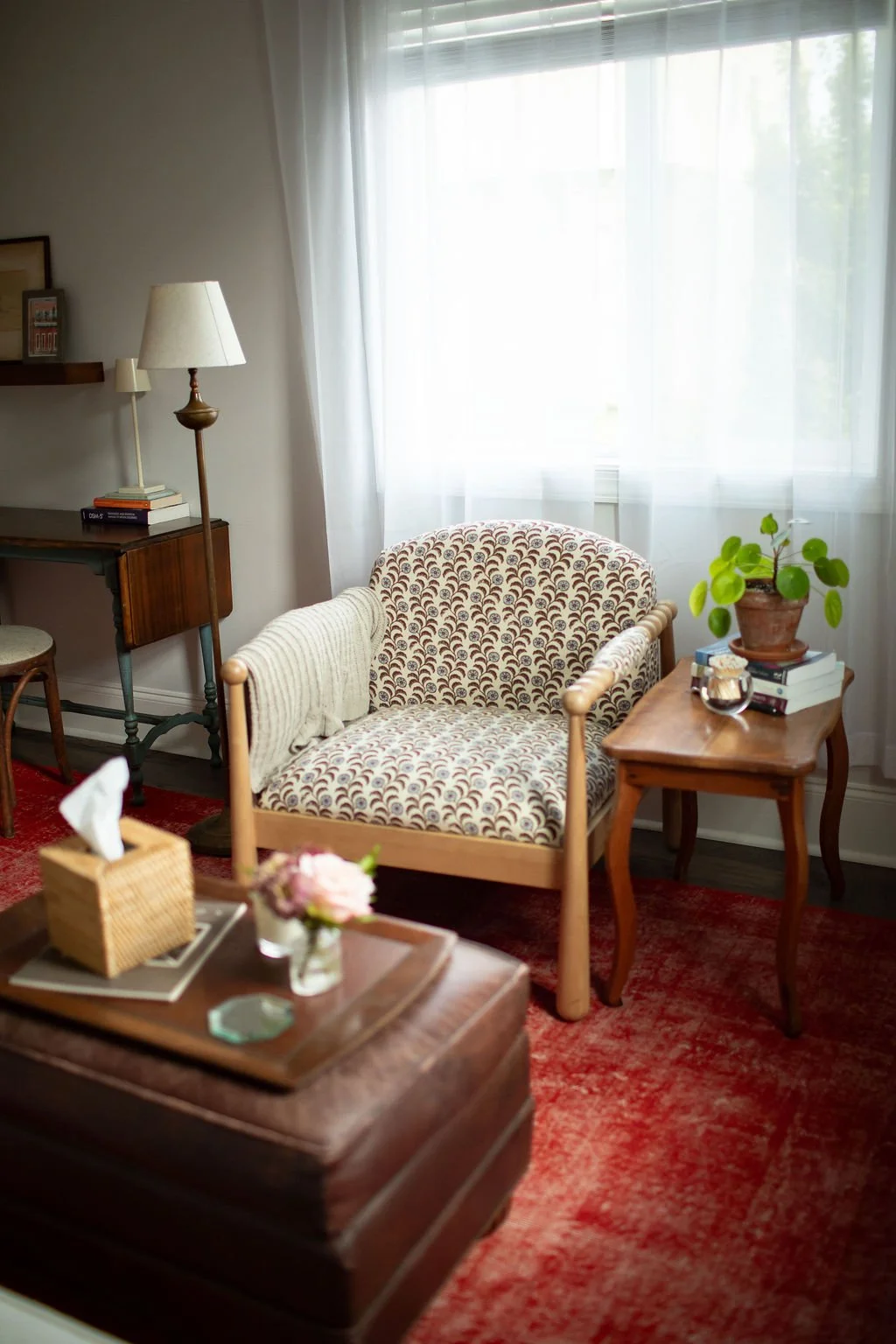A cozy therapy office corner with a patterned armchair, a small wooden side table with a potted plant, books, and decorative items, a window with sheer curtains, vintage-style lamp, and a red rug on the floor.