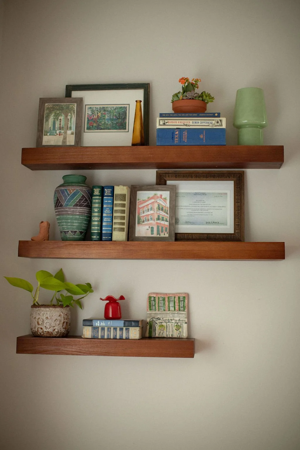 Three wooden wall shelves with books, framed pictures, vases, a plant, and decorative objects against a plain wall.