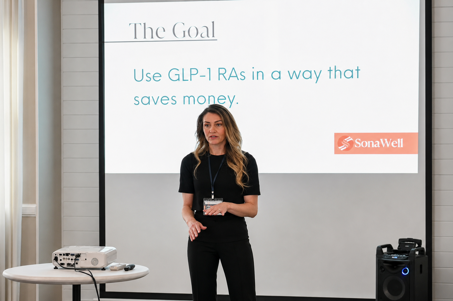 A woman giving a presentation in front of a large screen that displays the title 'The Goal' and the subtitle 'Use GLP-1 RAs in a way that saves money.' There is a table with a projector and a remote control, and a speaker on the right side of the image.