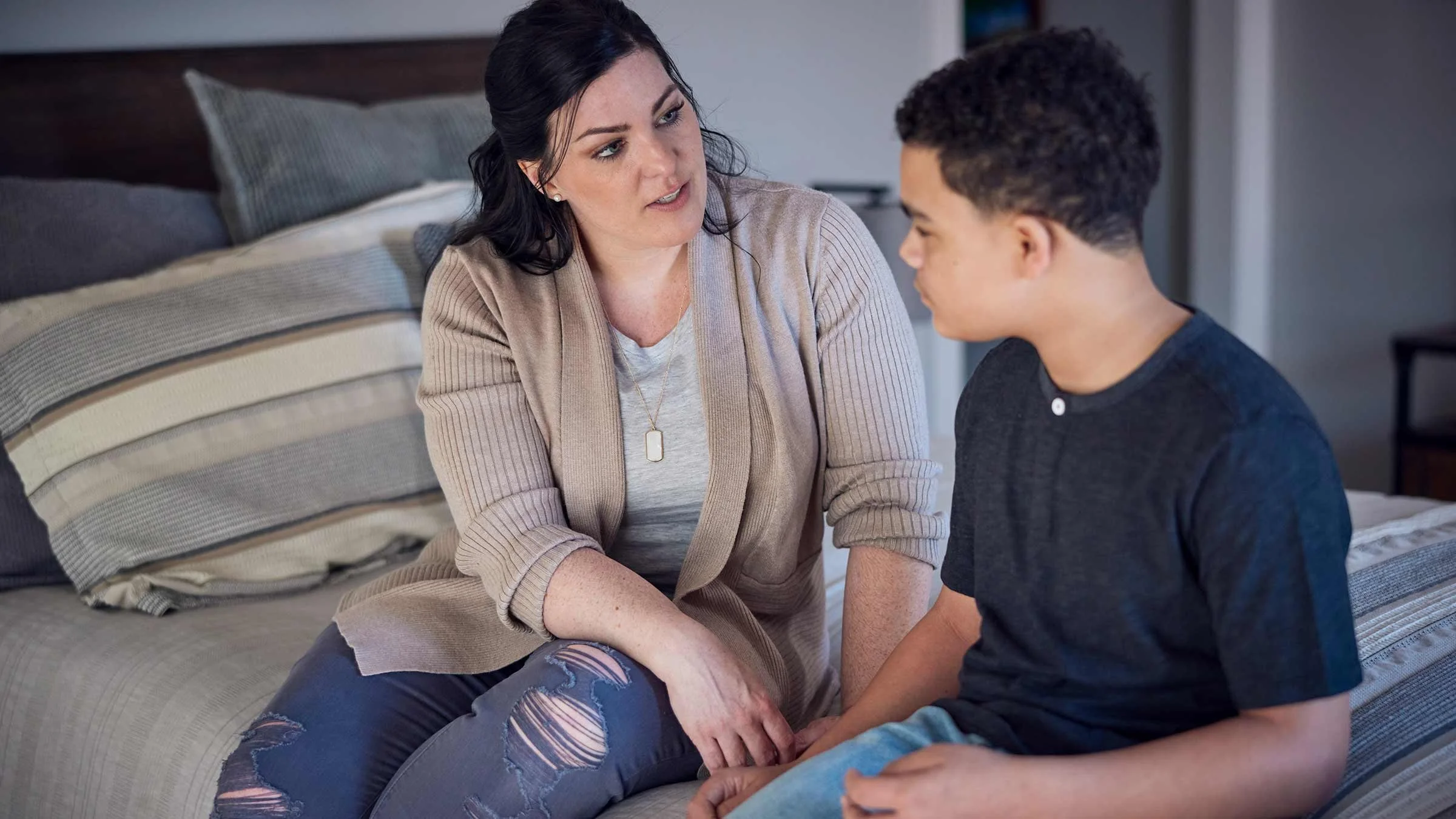 A woman and a young man sitting on a bed engaged in a serious conversation indoors.