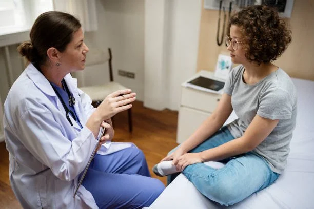 A female doctor consulting with a young female patient seated on a hospital bed in a medical office.