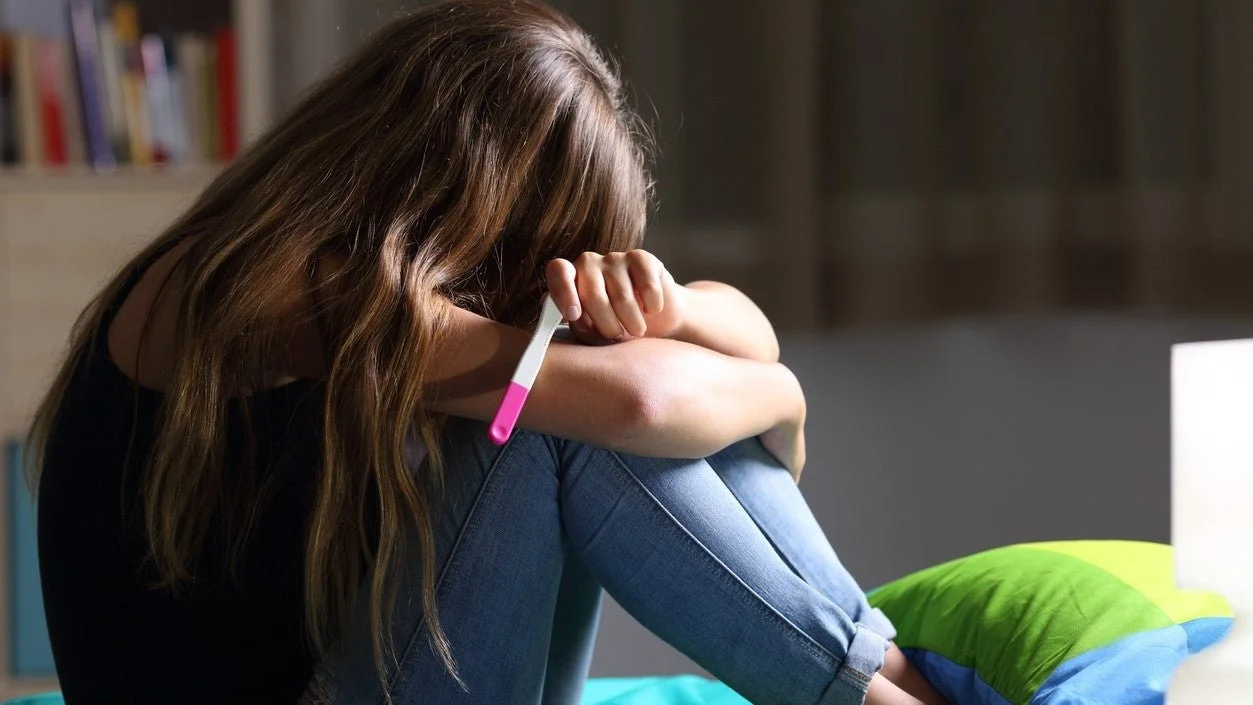 A girl sitting on her bed holding a pregnancy test with a positive result, looking distressed in a dimly lit room.