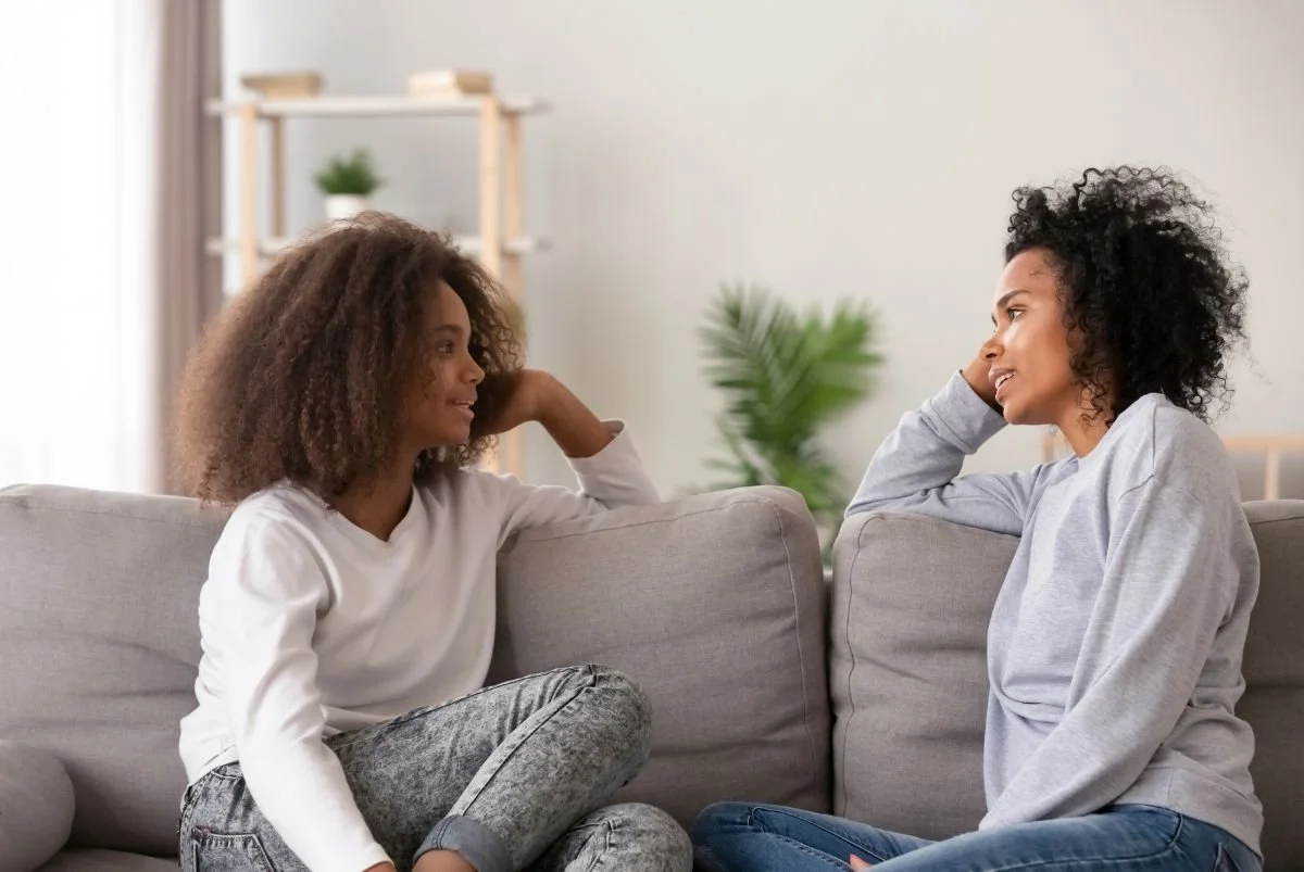 Two women, a mother and daughter, sitting on a gray couch and talking in a well-lit living room with a plant in the background.