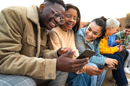 Group of diverse young people sitting outdoors, looking at their phones and smiling.