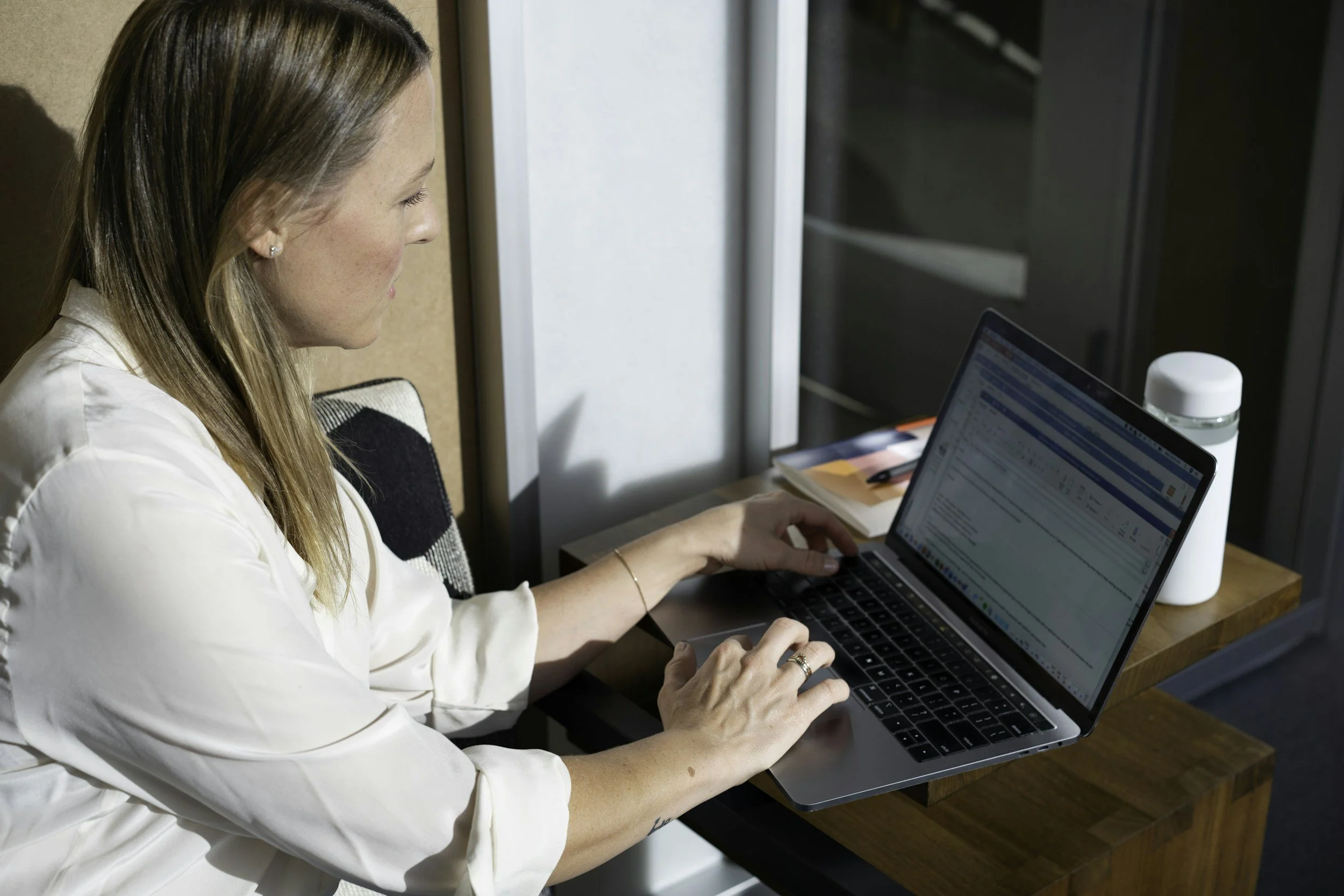 A woman working on a laptop at a desk near a window, with a water bottle and notebooks beside her.