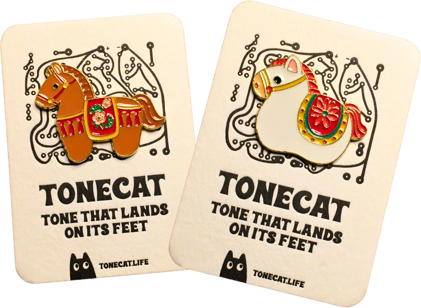 Two enamel pins featuring stylized horses on gift cards with black and white abstract background designs and message about the tone that lands on its feet. One pin is on a brown horse with a red and green saddle, and the other is on a white horse with similar decorative elements.