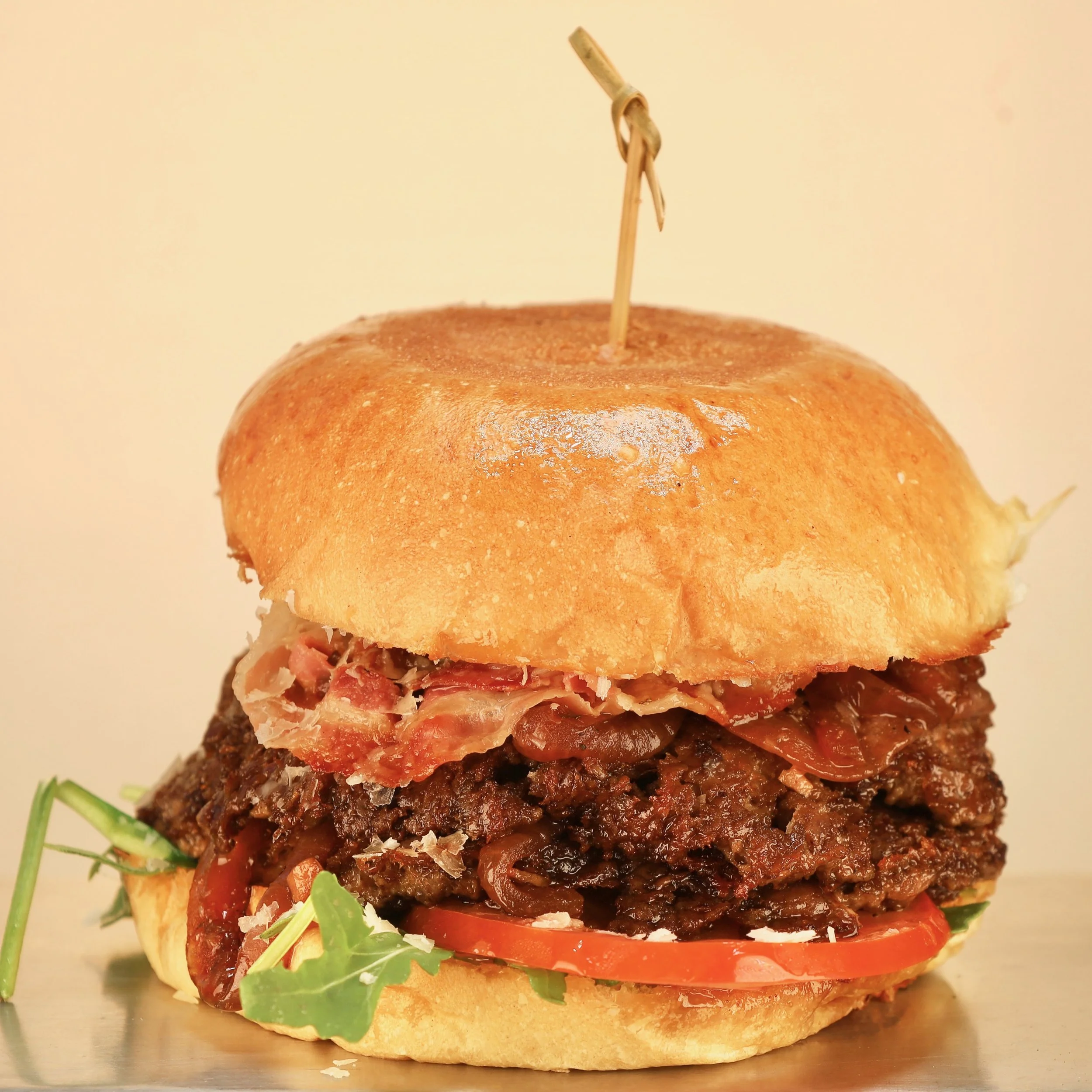 Close-up of a gourmet hamburger sandwich with tomato, arugula, caramelized onions, shredded cheese, crispy bacon, and a thick beef patty inside a toasted bun with a toothpick on top.