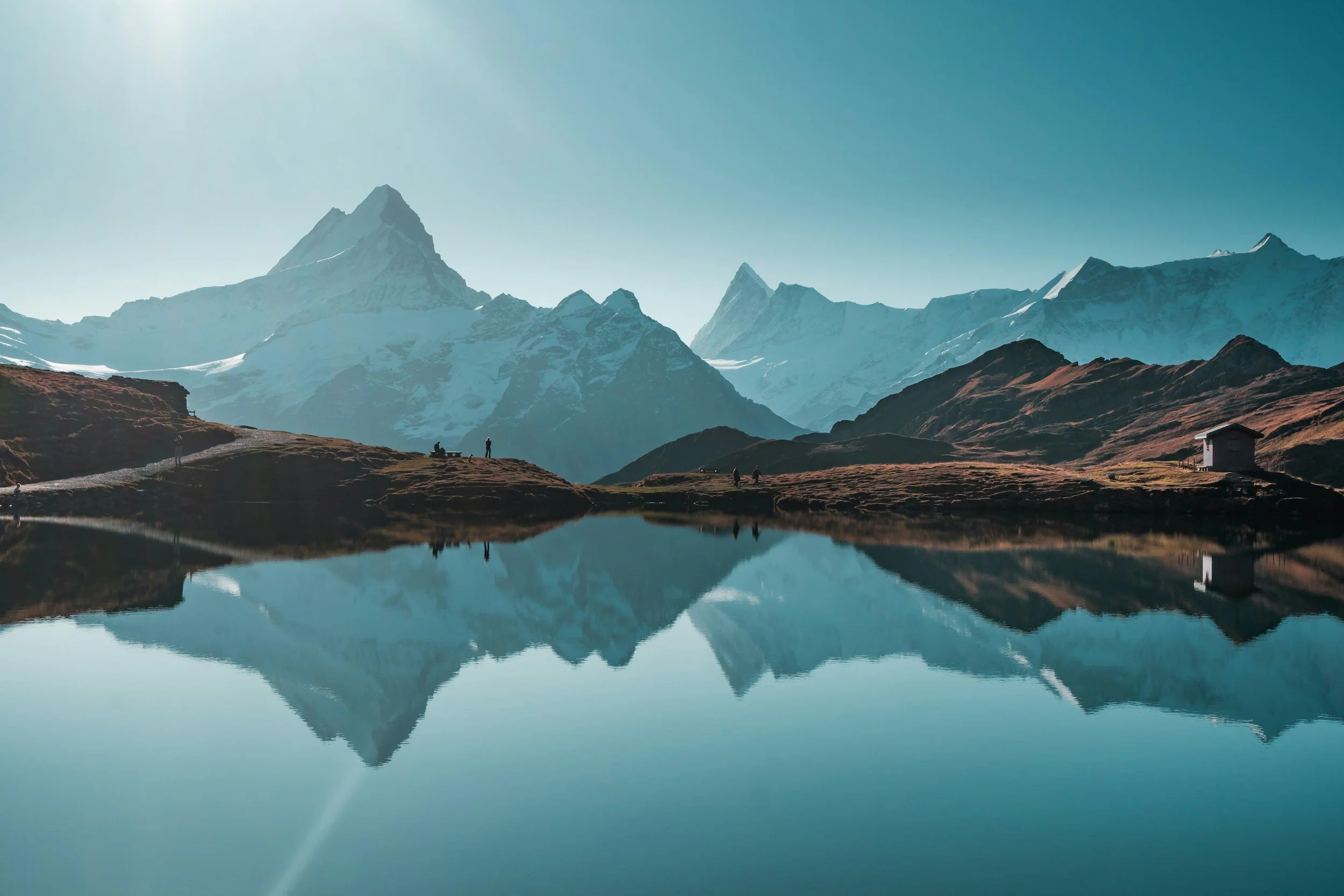 Snow-capped mountains reflected in a calm lake with a few small structures and people on grassy land.
