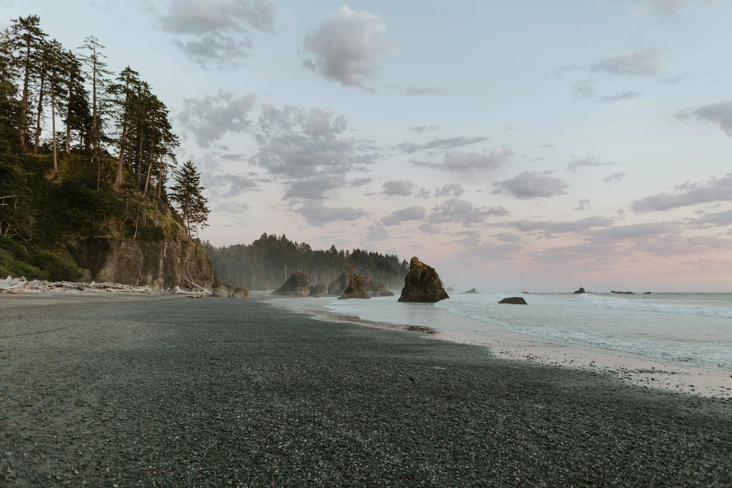 A peaceful black sand beach with large rock formations and a forested cliff on the left under a partly cloudy sky.